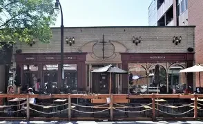 An exterior view of a restaurant featuring outdoor seating with umbrellas, surrounded by trees and buildings.