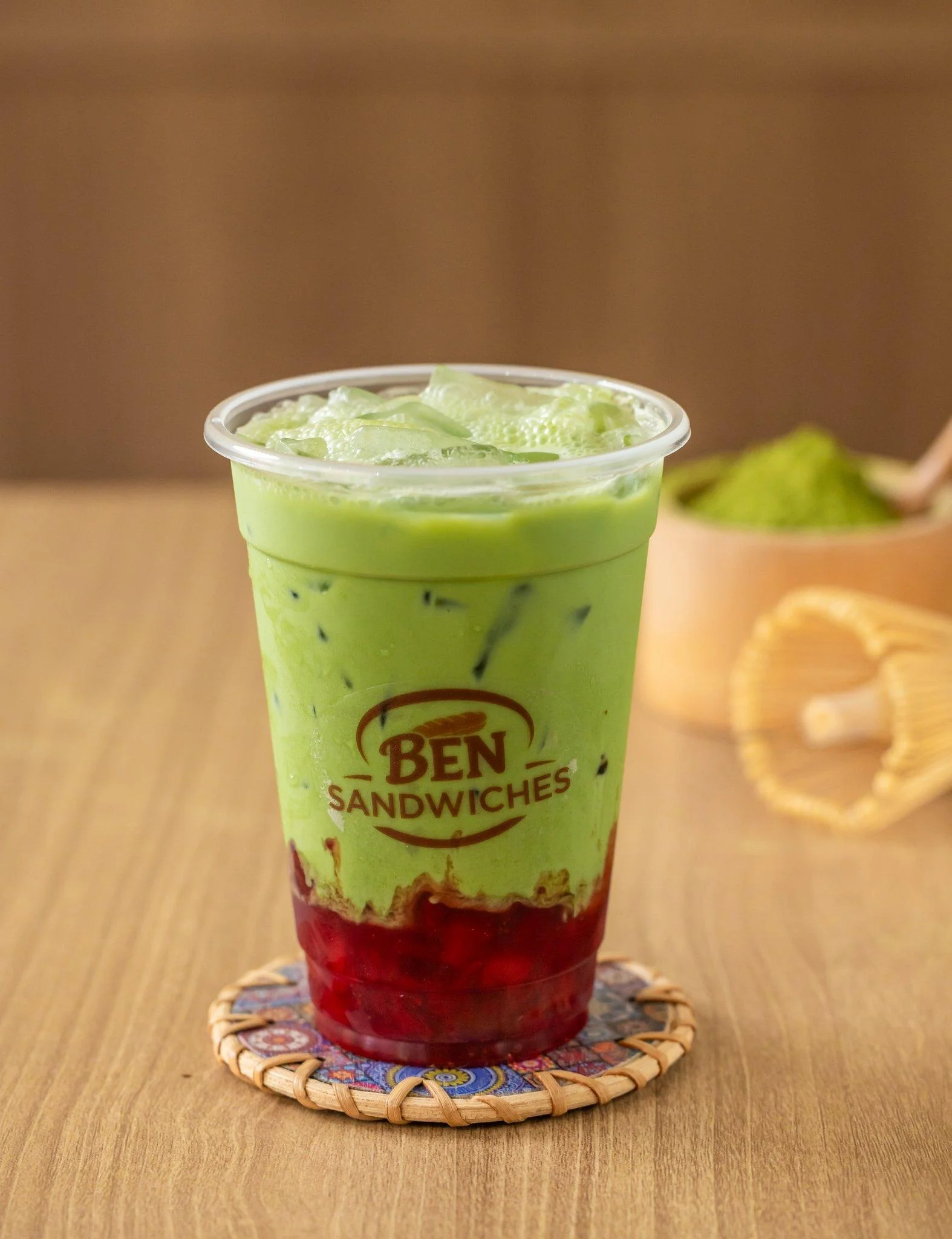 A layered iced drink featuring green and red components, topped with ice cubes. A small bowl of matcha powder is visible nearby.