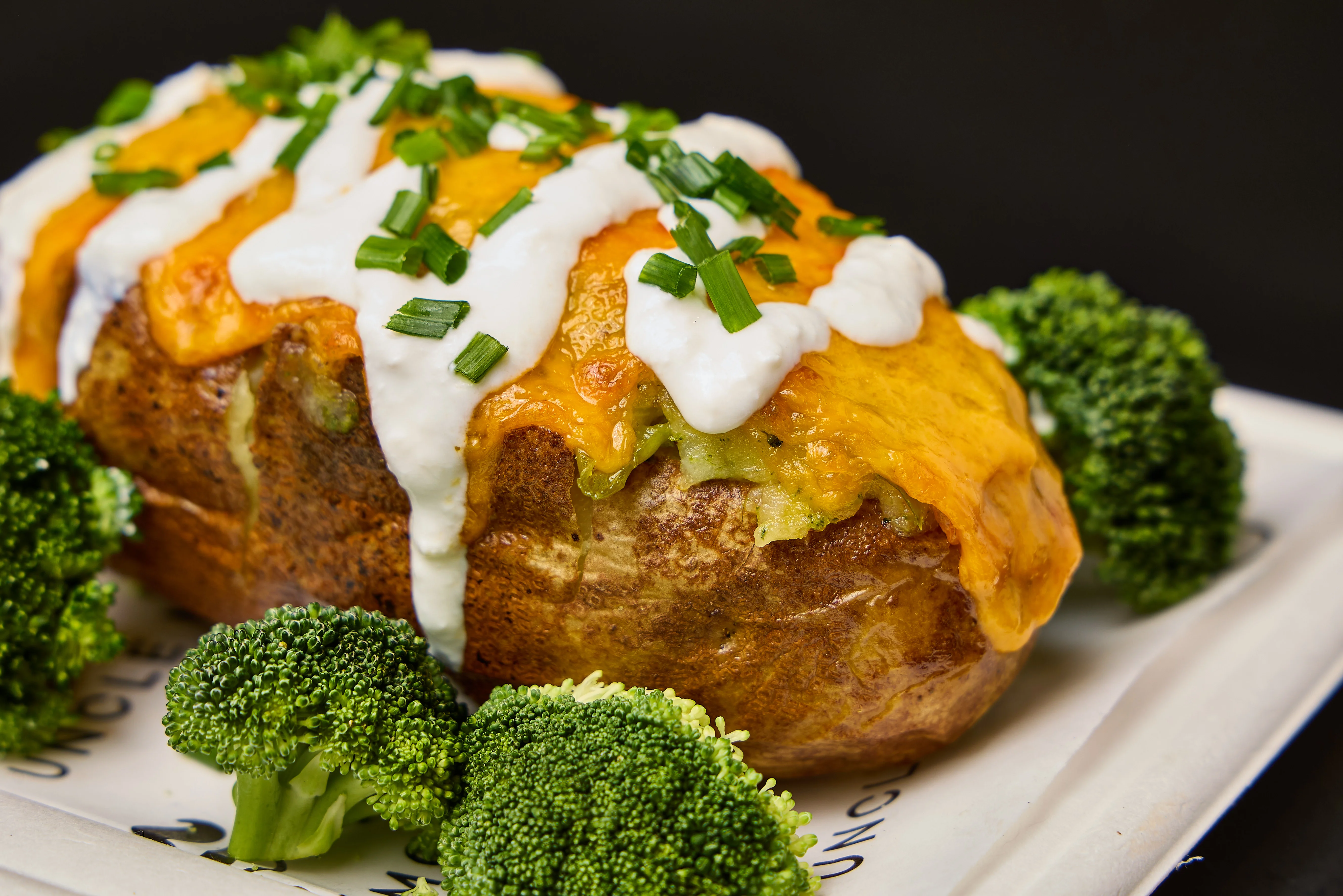 A baked potato topped with cheese, sour cream, and chives, served with broccoli on the side.