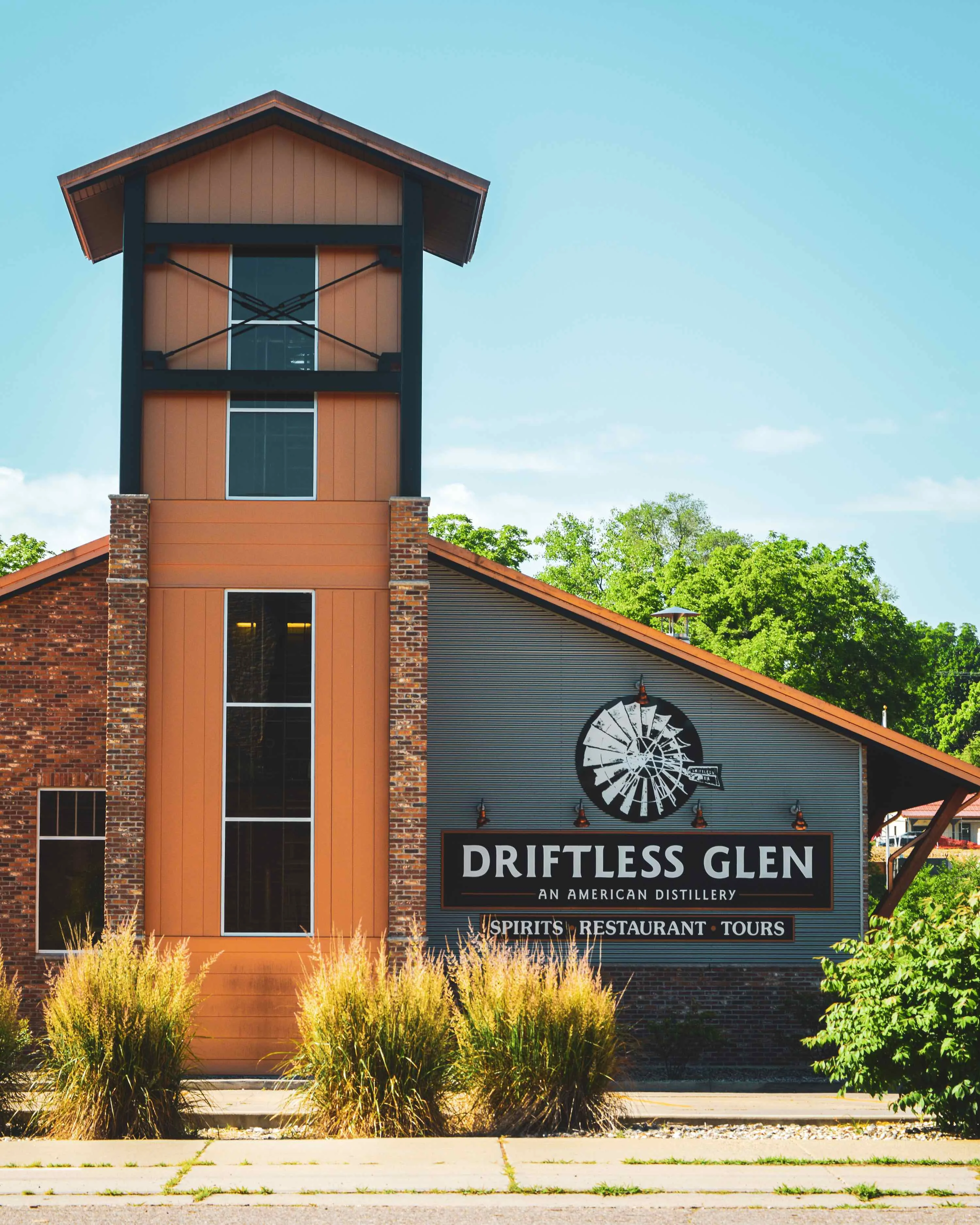 This image is of the outside of the Driftless Glen building, taken from a distance, looking over the parking lot and bushes to see the front of the distillery