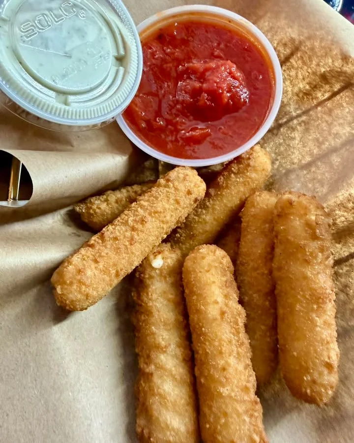 A serving of crispy cheese sticks accompanied by marinara sauce and a ranch-style dip, presented in a basket lined with paper.