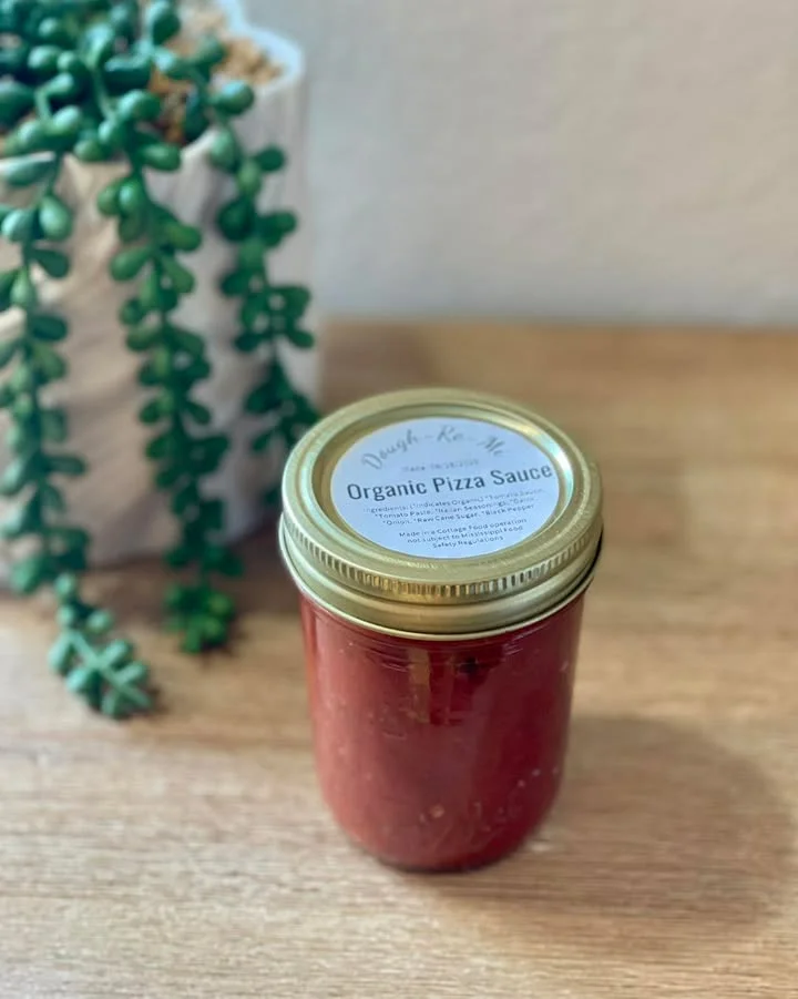 A jar of organic pizza sauce sitting on a wooden surface, next to a green plant.