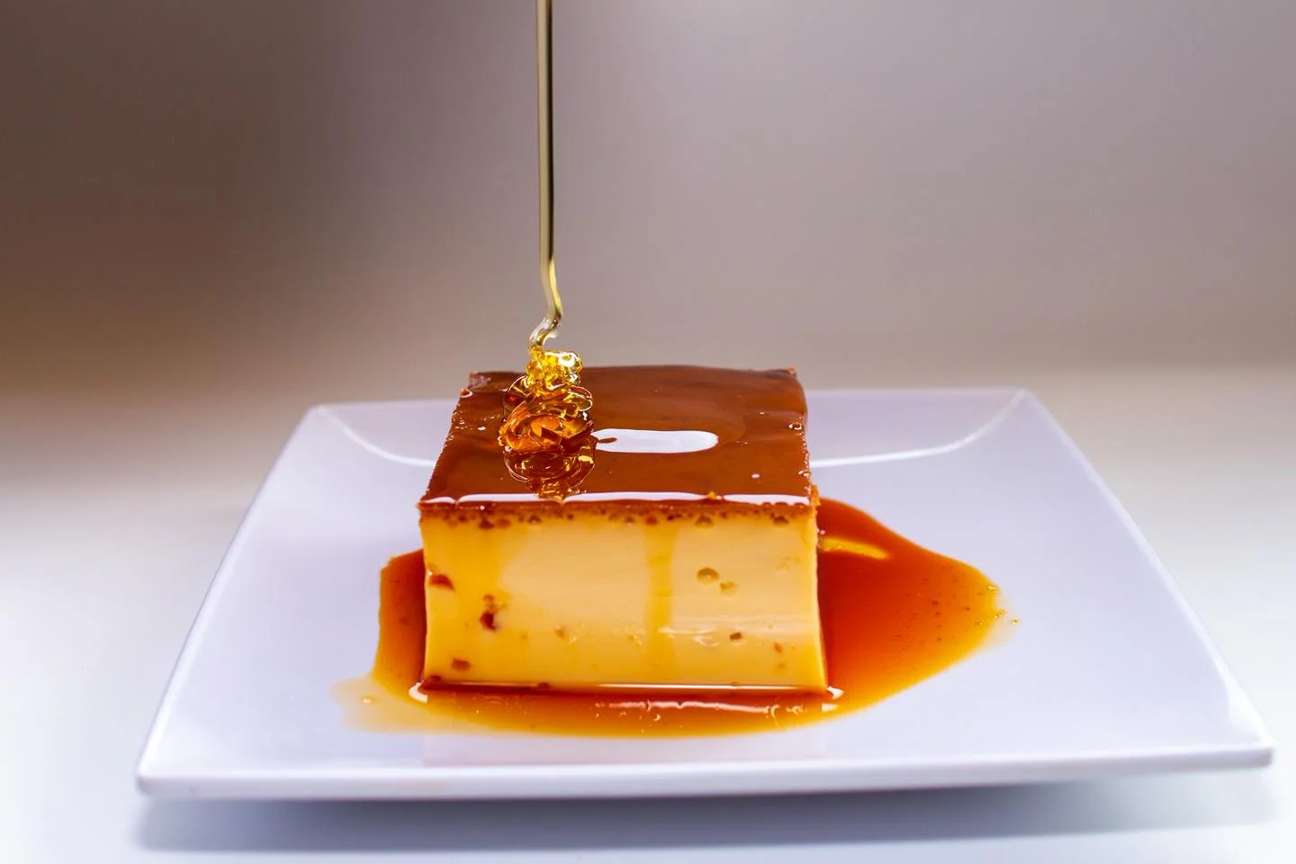 A creamy caramel dessert topped with a golden syrup drizzling from above, presented on a white plate.