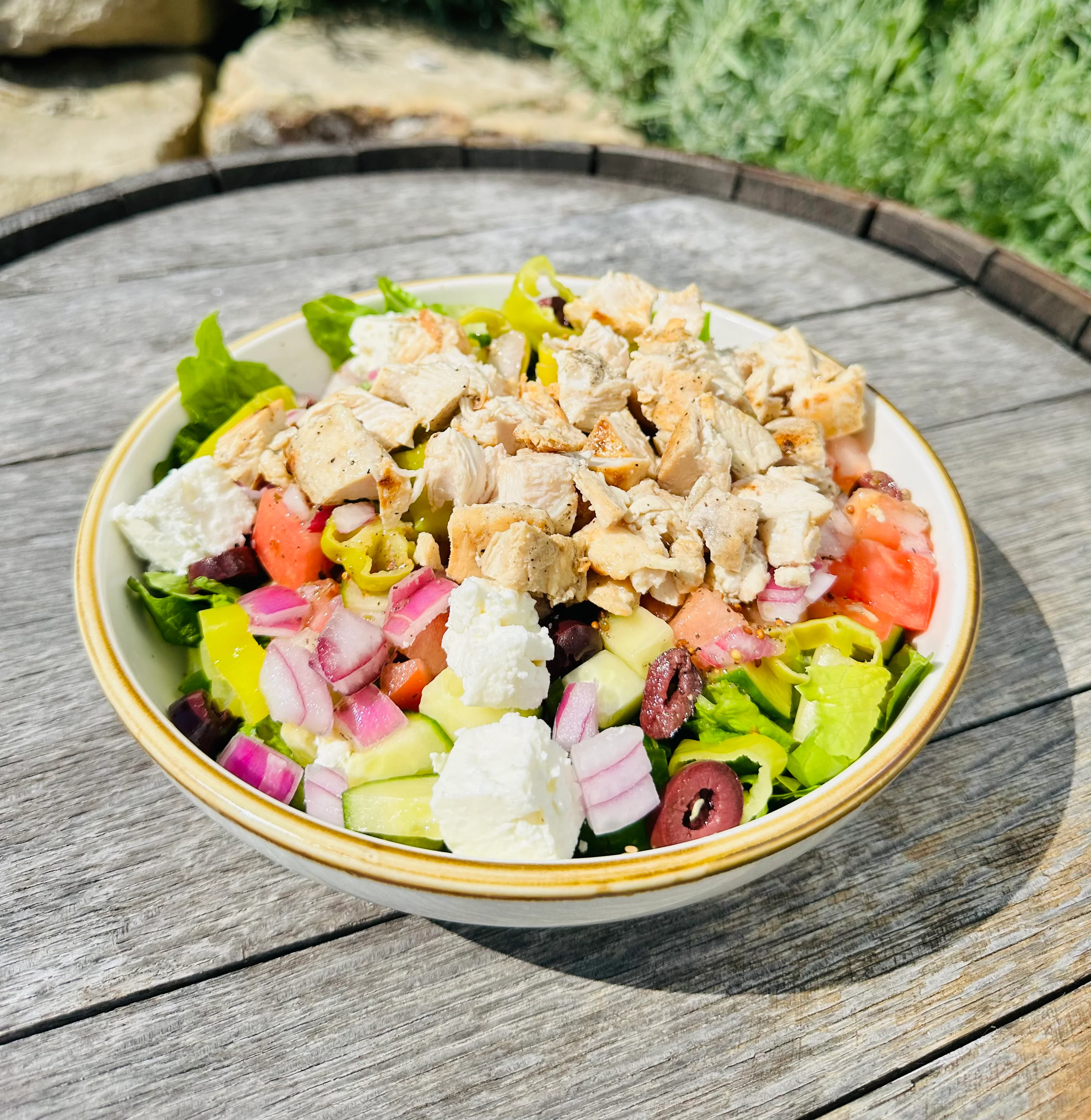 A vibrant salad bowl filled with mixed greens, chopped tomatoes, cucumbers, onions, peppers, and topped with grilled chicken and feta cheese, placed on a wooden table.