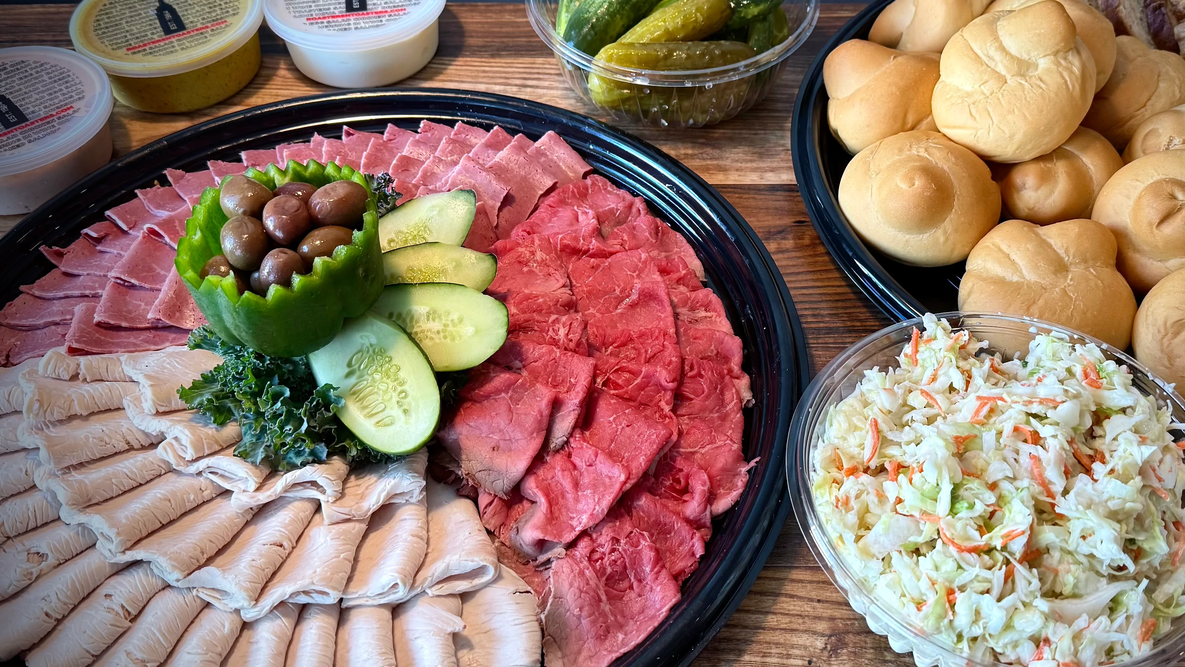Deli catering spread with sliced meats, bread rolls, and coleslaw