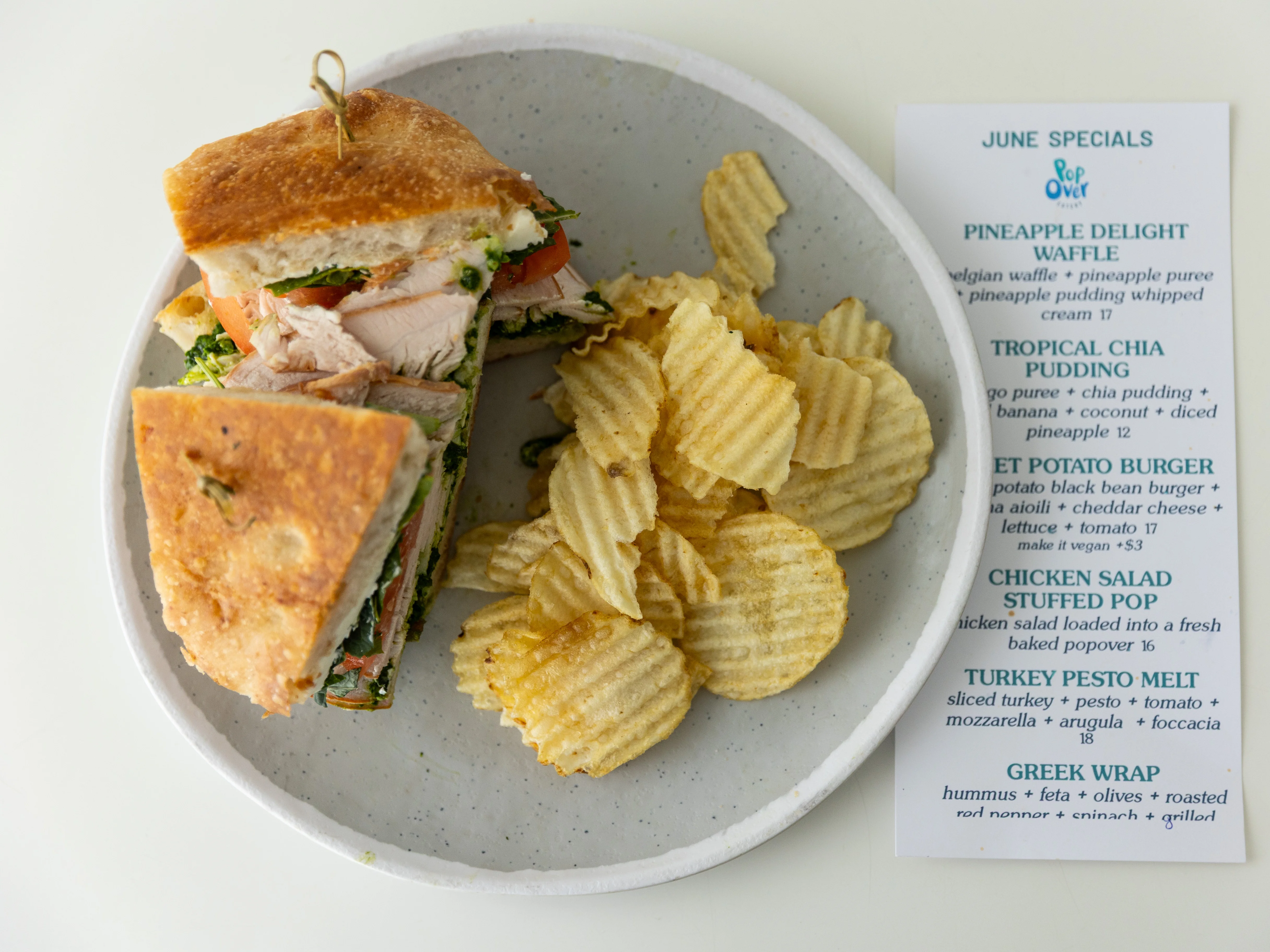 A sandwich filled with turkey, greens, and tomato, accompanied by a side of potato chips. A menu is visible in the background.