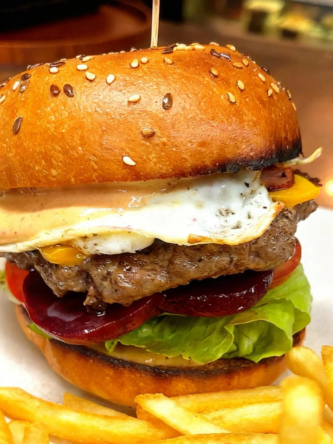 A juicy burger featuring a beef patty, cheese, lettuce, tomatoes, and a fried egg, all nestled in a sesame seed bun. Accompanied by fries.