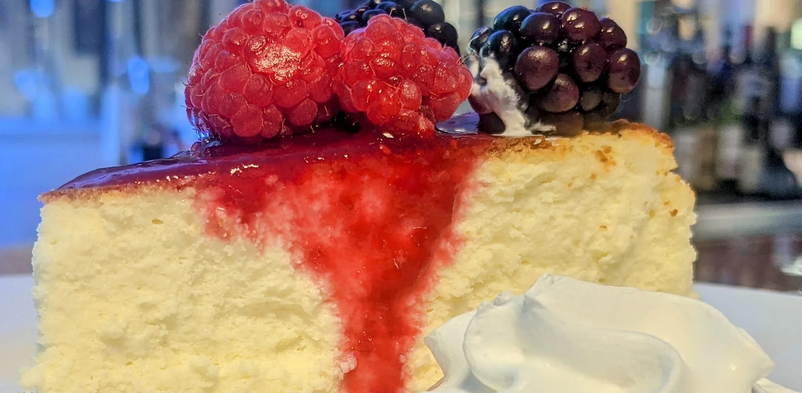 A slice of cheesecake topped with raspberry and blackberry, served with a dollop of whipped cream on a white plate.