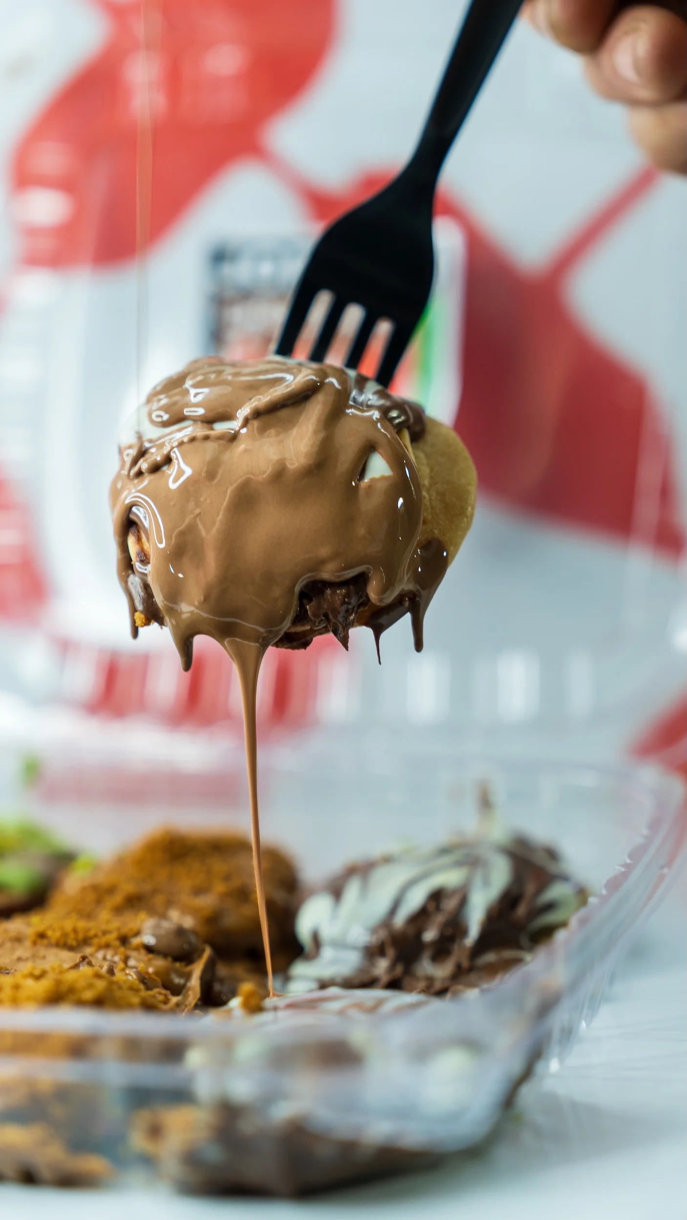A fork lifting a dessert topped with melted chocolate, drizzling down, from a clear plastic container filled with various sweet treats.