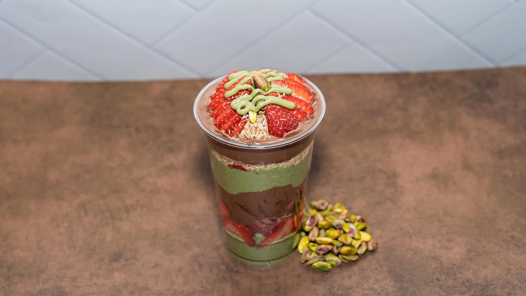 A layered dessert in a clear cup featuring chocolate and green layers, topped with strawberries and decorative drizzle. Beside it are some pistachios.