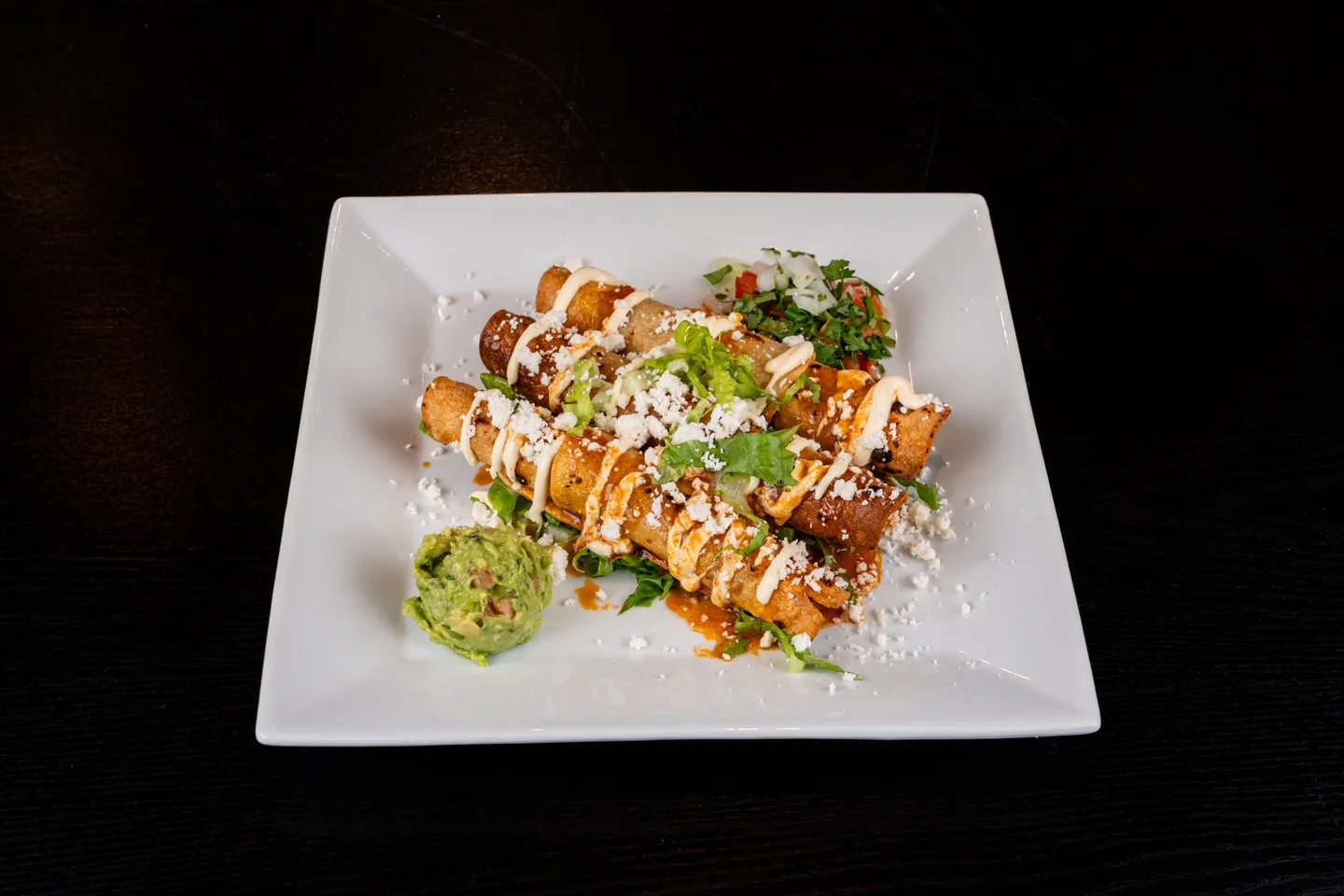 A plate of taquitos topped with crema, cheese, and fresh lettuce, alongside a scoop of guacamole.