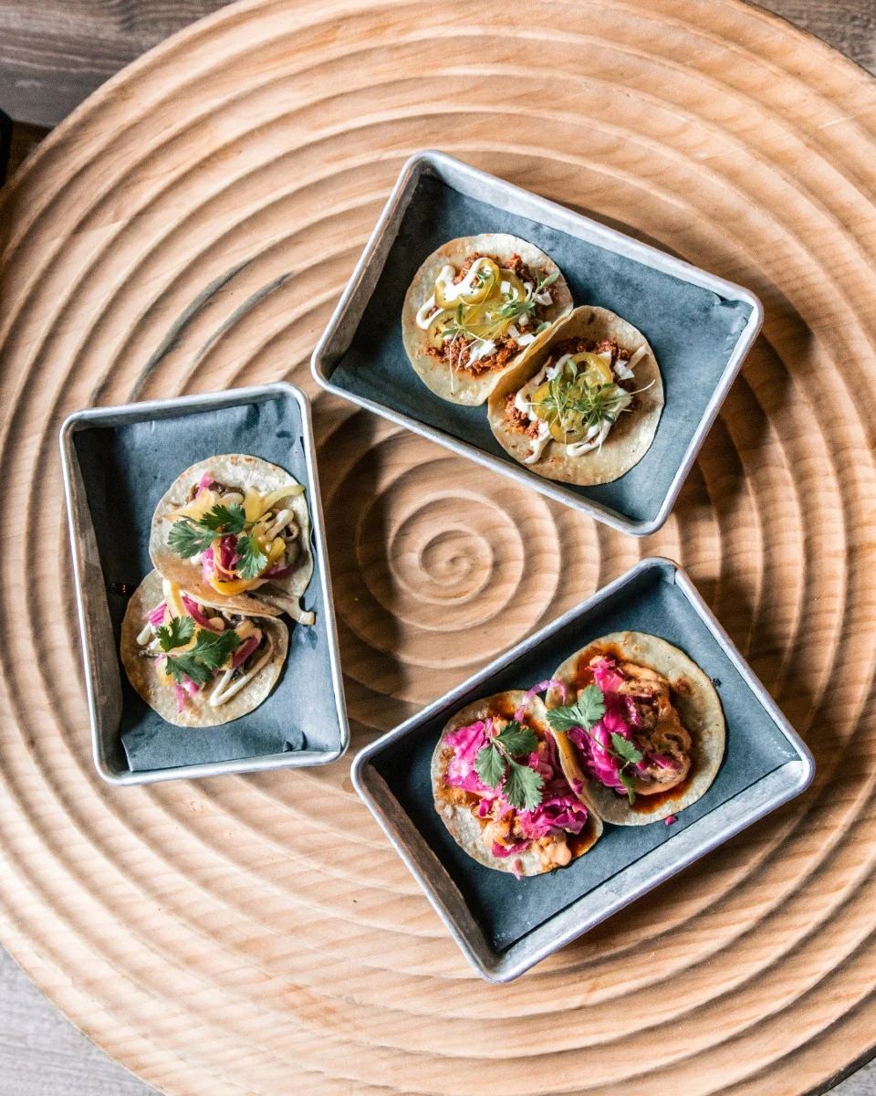 A selection of three tacos served on metal trays, showcasing various fillings and garnished with fresh herbs on a decorative wooden table.