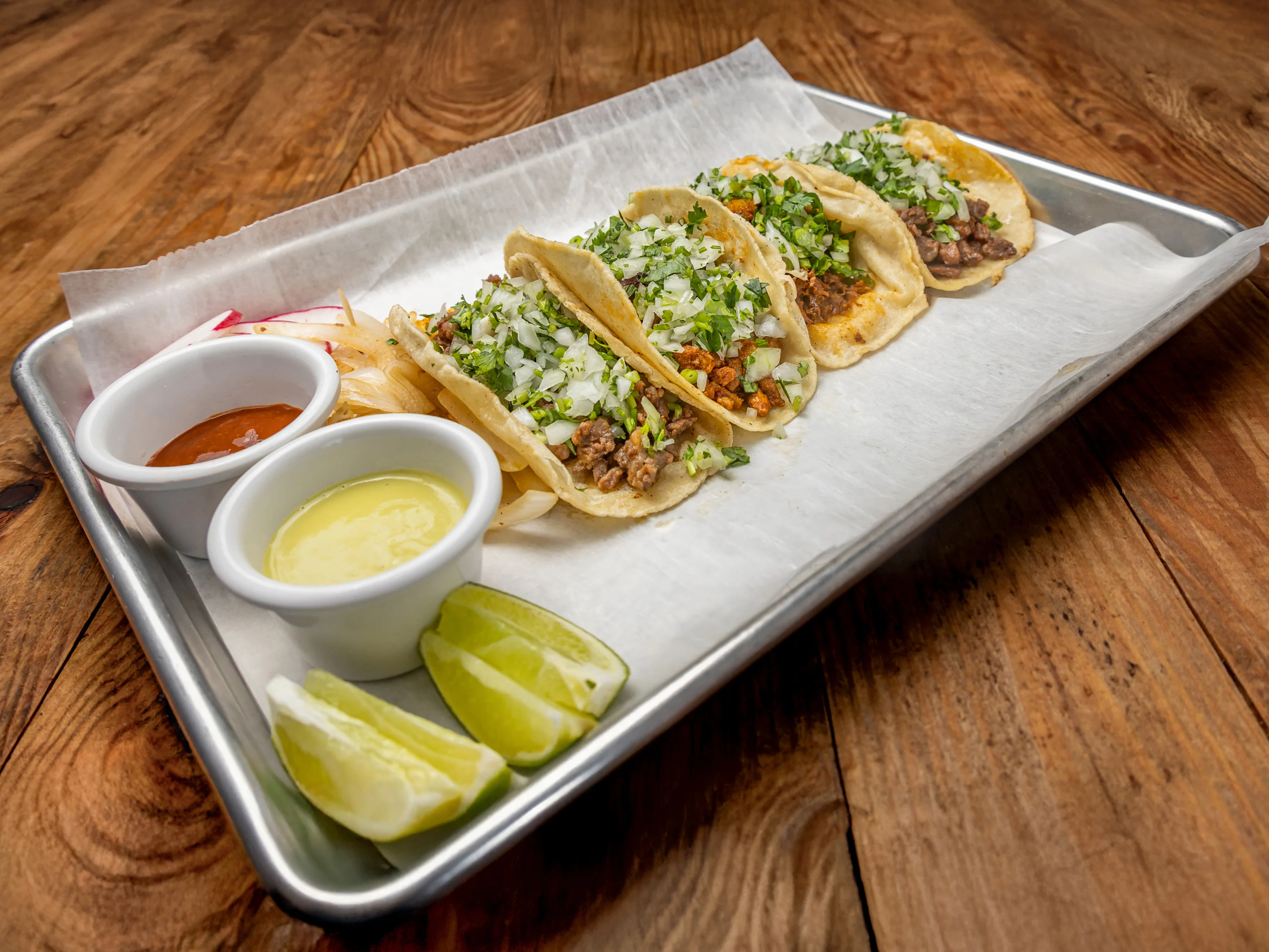 Three tacos loaded with slaw, cheese, and fresh salsa, served with lime wedges and a side of dipping sauce.