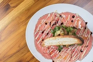 A plate of thinly sliced meat arranged artfully with salad and drizzled with sauce, accompanied by a slice of bread.