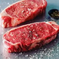 Two raw ribeye steaks seasoned with salt and pepper on a metal surface.