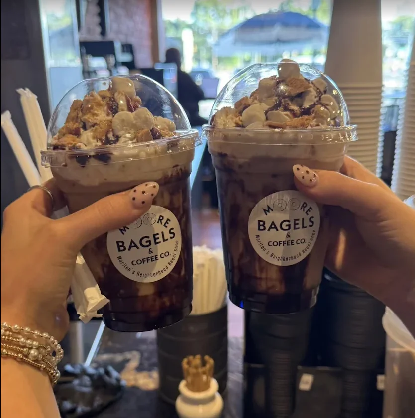 A hand holding an iced coffee beverage with a chocolate drizzle on top, set against a textured dark surface.
