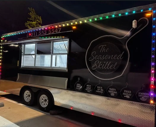 A food truck decorated with colorful lights, showcasing its name 'The Seasoned Skillet' on the side. The window is open, revealing the interior.