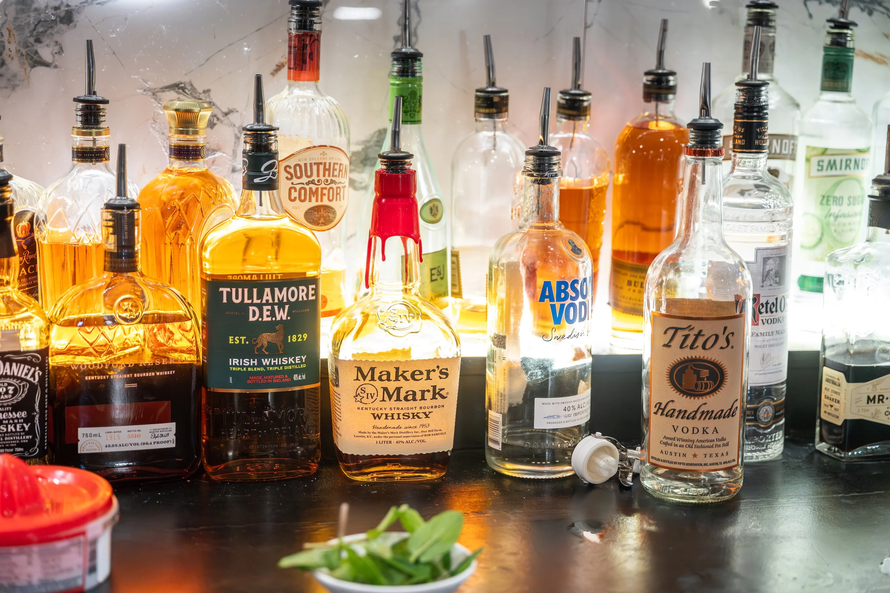 A backlit bar shelf displays various liquor bottles, including whiskey and vodka, with pour spouts. The scene is vibrant, suggesting a lively bar atmosphere.