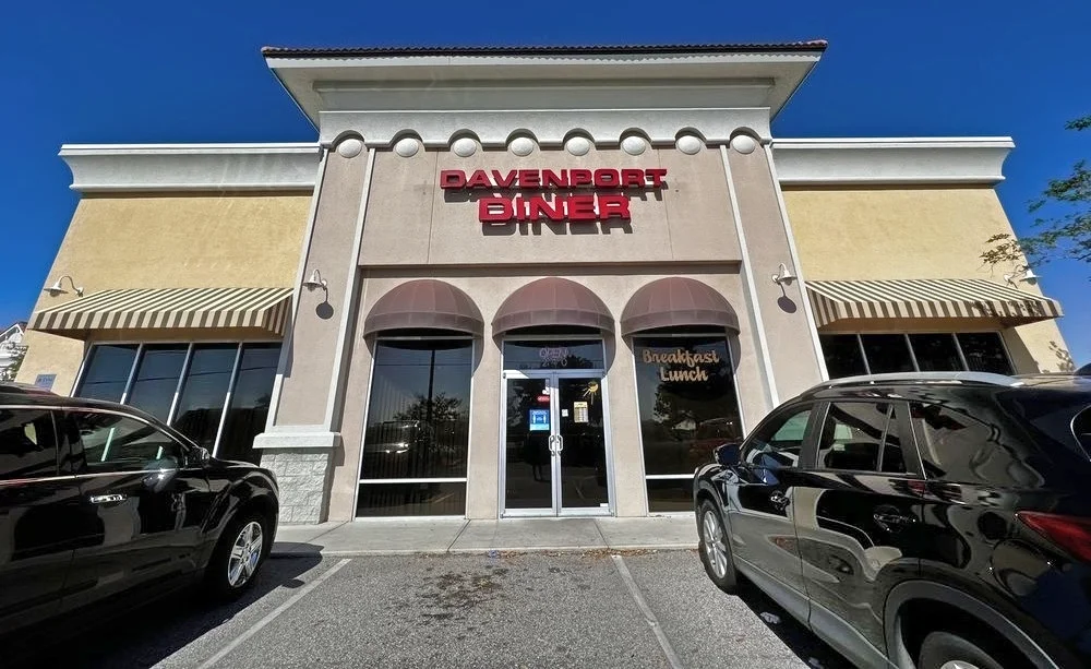 Front view of Davenport Diner with a beige facade, red 'Davenport Diner' sign, and large windows, flanked by parked cars under a clear blue sky.