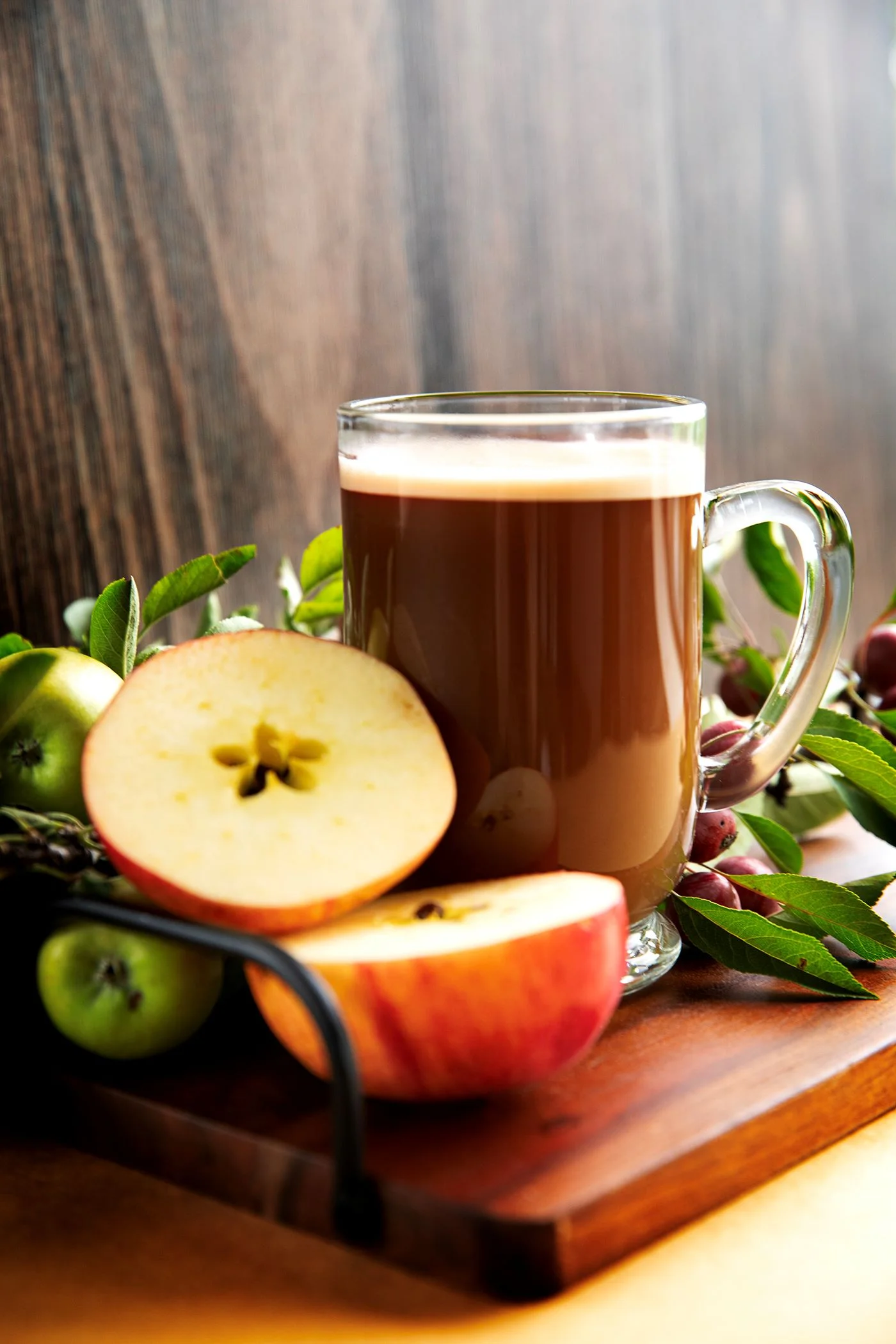 A glass mug filled with warm caramel-colored apple cider sits on a wooden board, surrounded by fresh apples and green leaves, conveying a cozy autumn feel.