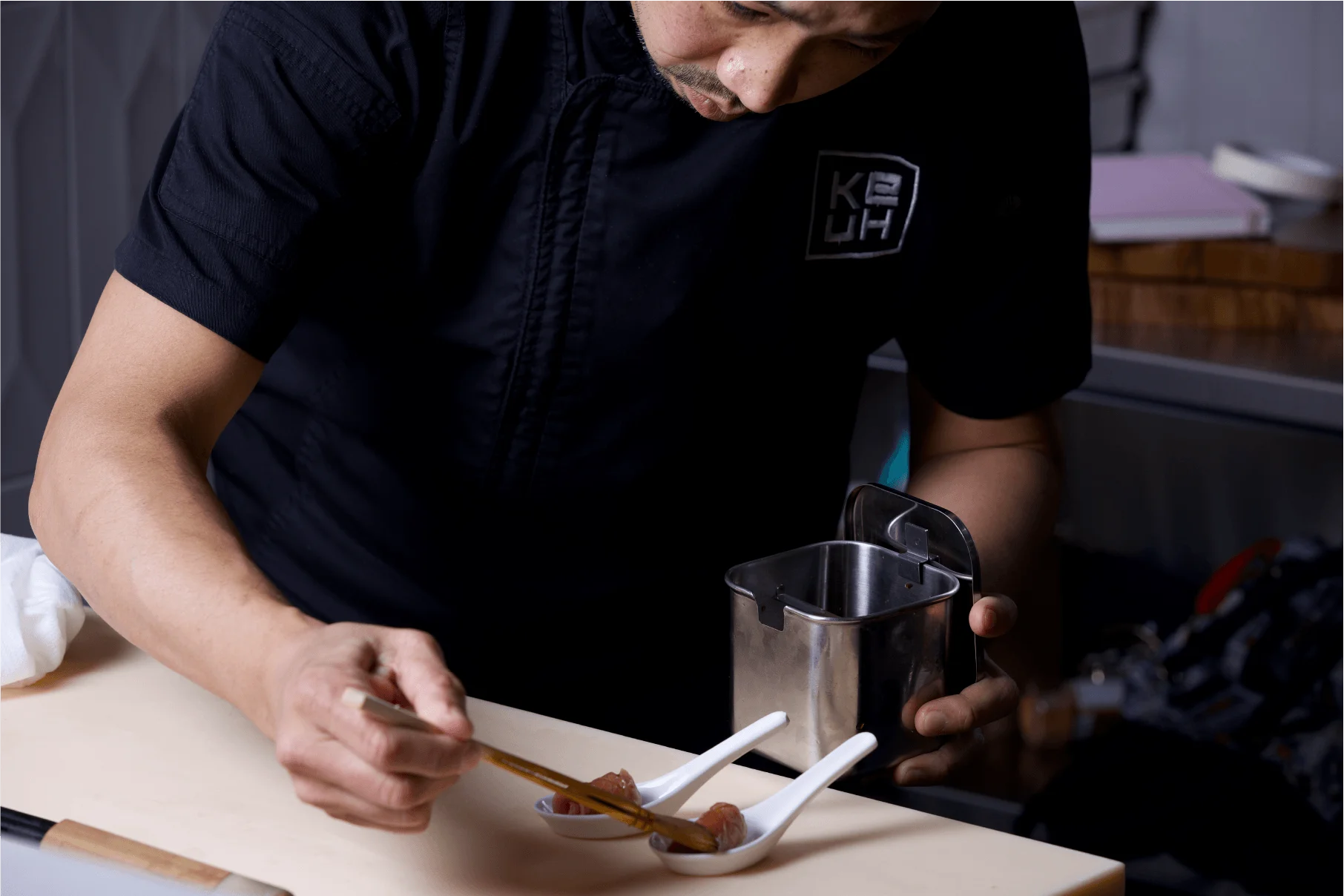 A chef wearing a branded Ke-uH uniform uses a brush to apply sauce from a stainless steel container onto small appetizers served in individual white ceramic spoons.