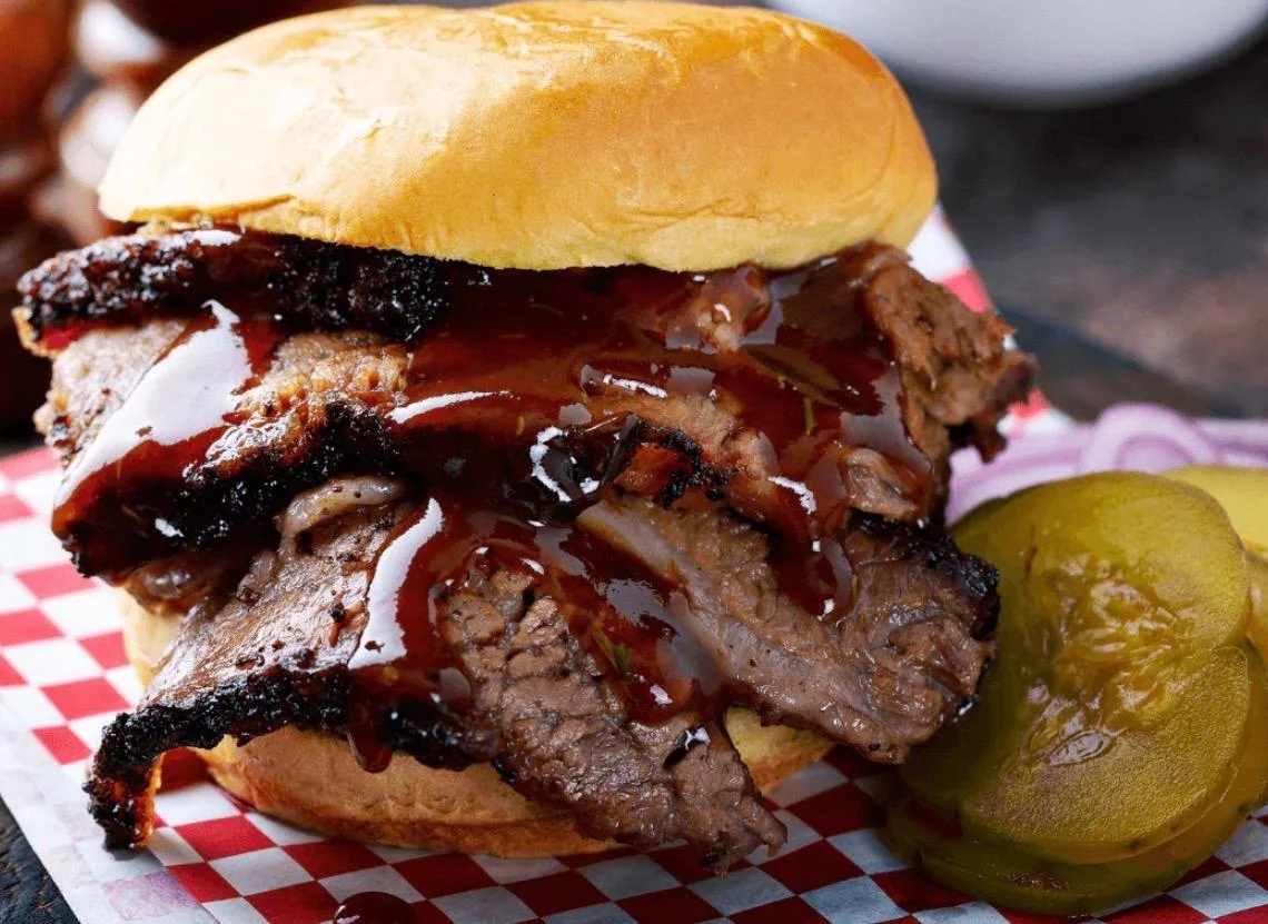 A hearty sandwich featuring a thick layer of smoked brisket topped with barbecue sauce, served on a soft bun alongside slices of pickles.