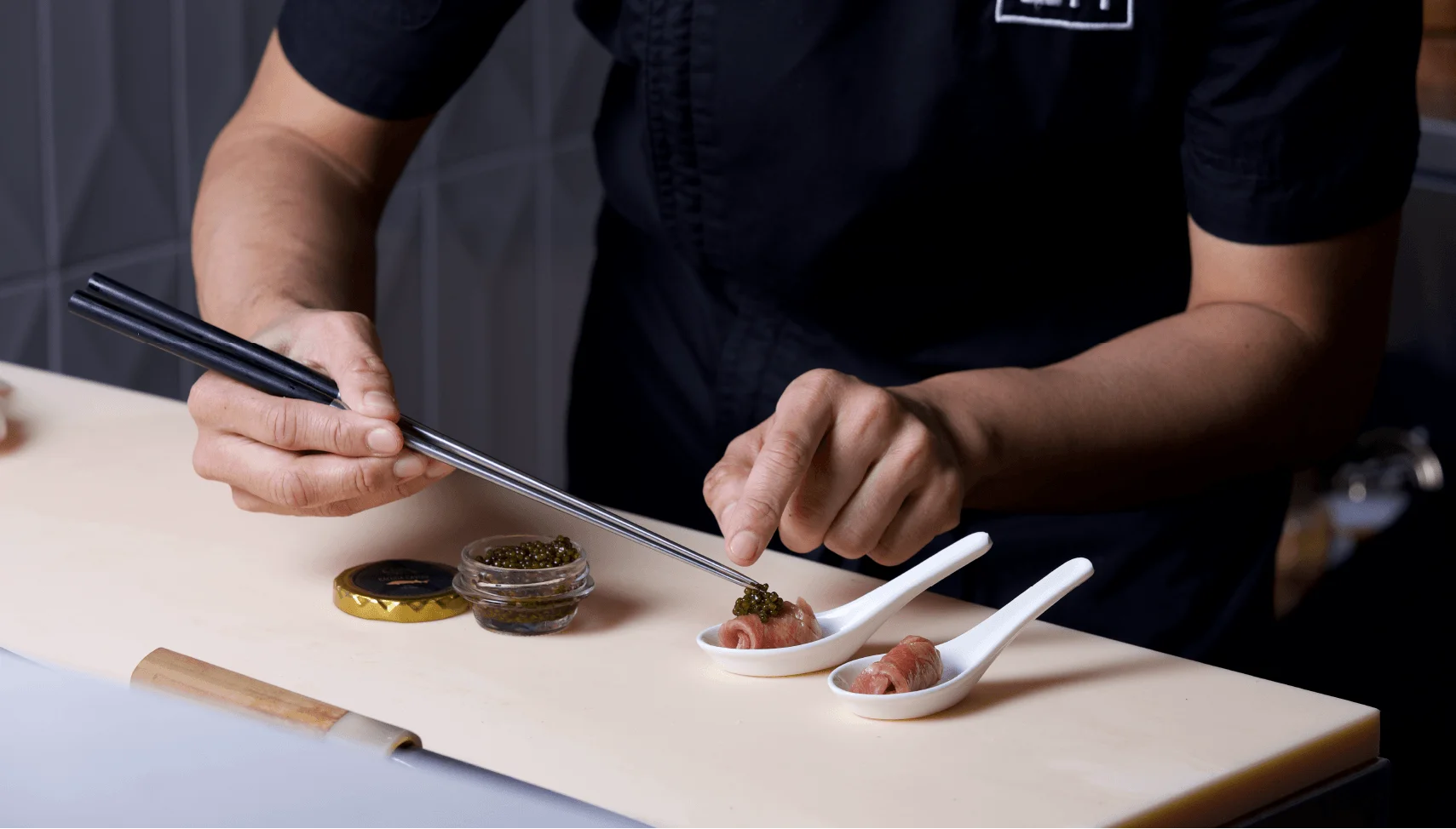 Ke-uH chef in a branded black uniform uses chopsticks to delicately place caviar onto rolled pieces of fish served in individual white tasting spoons.