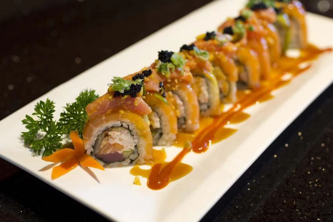 A beautifully arranged sushi platter showcasing various rolls topped with colorful garnishes and sauces on a white plate.