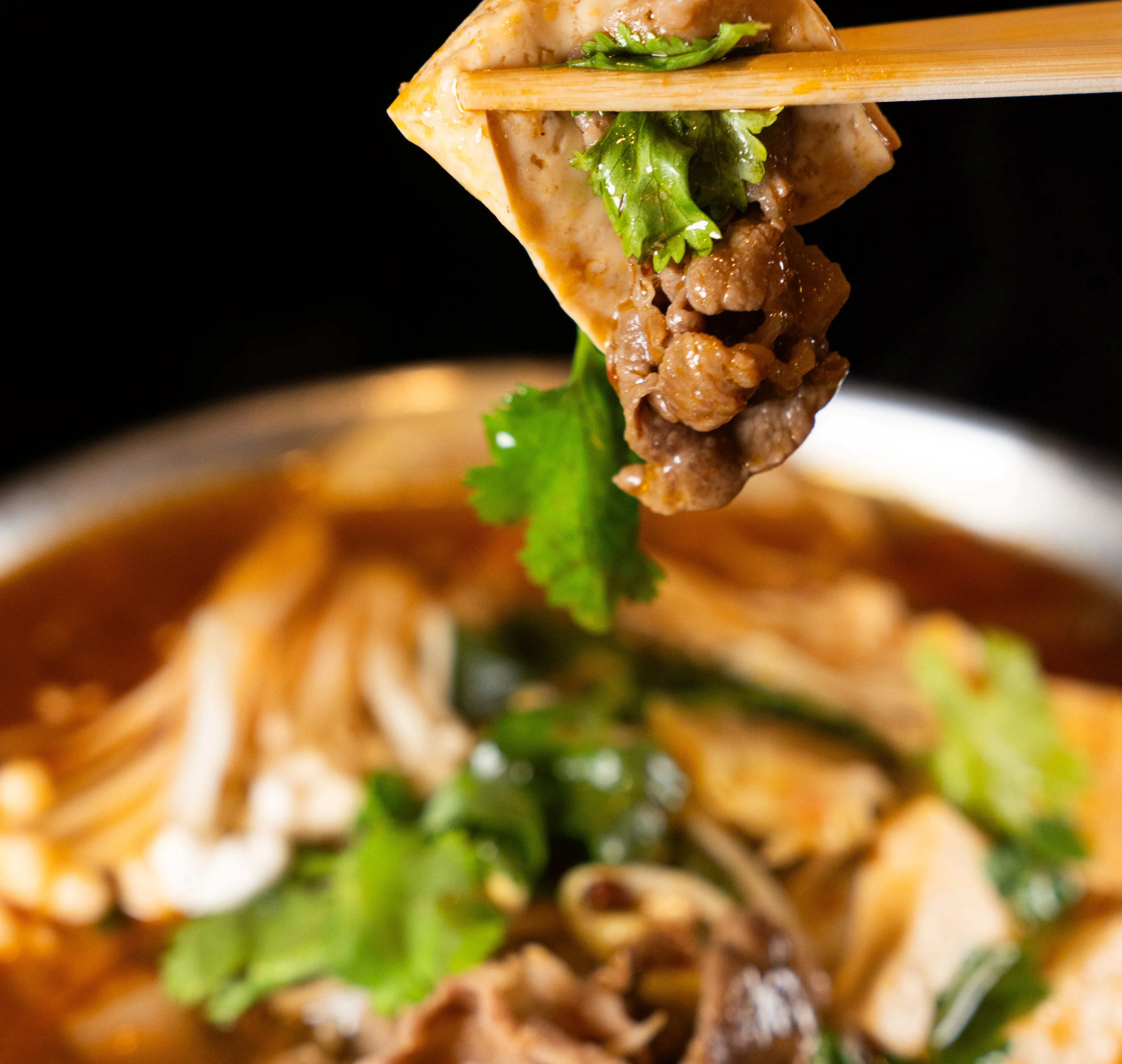 Chopsticks holding tofu and beef in spicy broth with herbs.