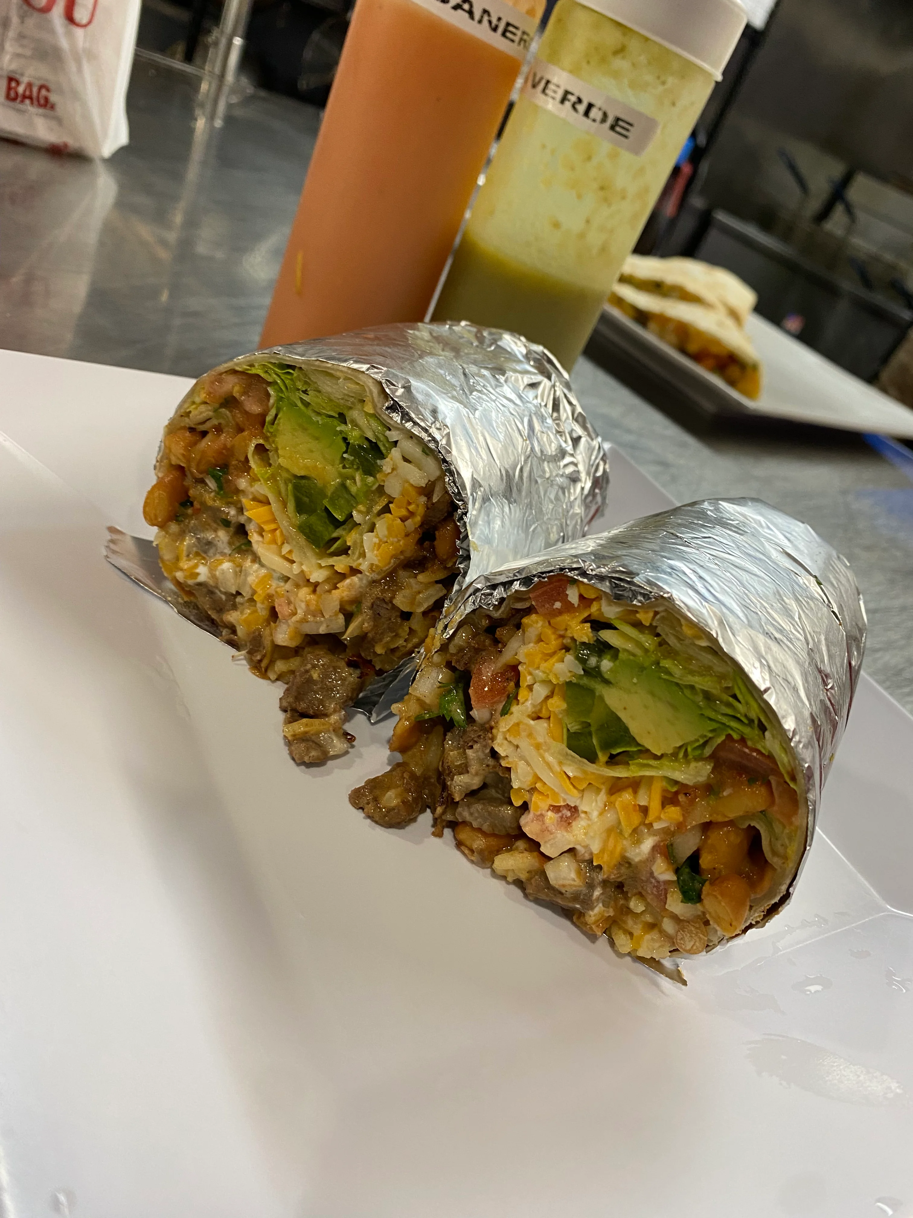 Two foil-wrapped burritos filled with rice, beans, lettuce, and meat, accompanied by colorful salsa bottles.