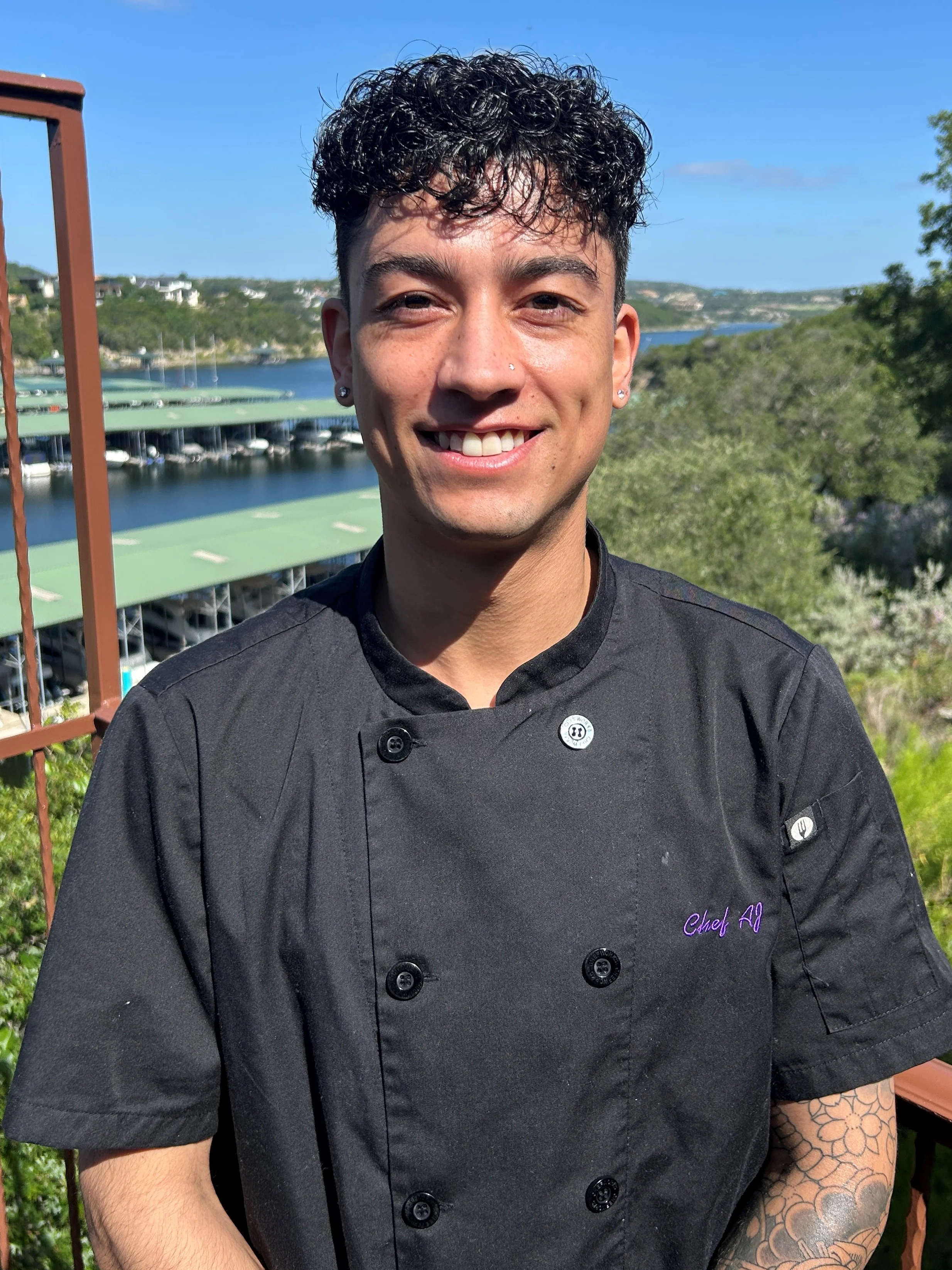 Chef in a black uniform smiling outdoors with a scenic view