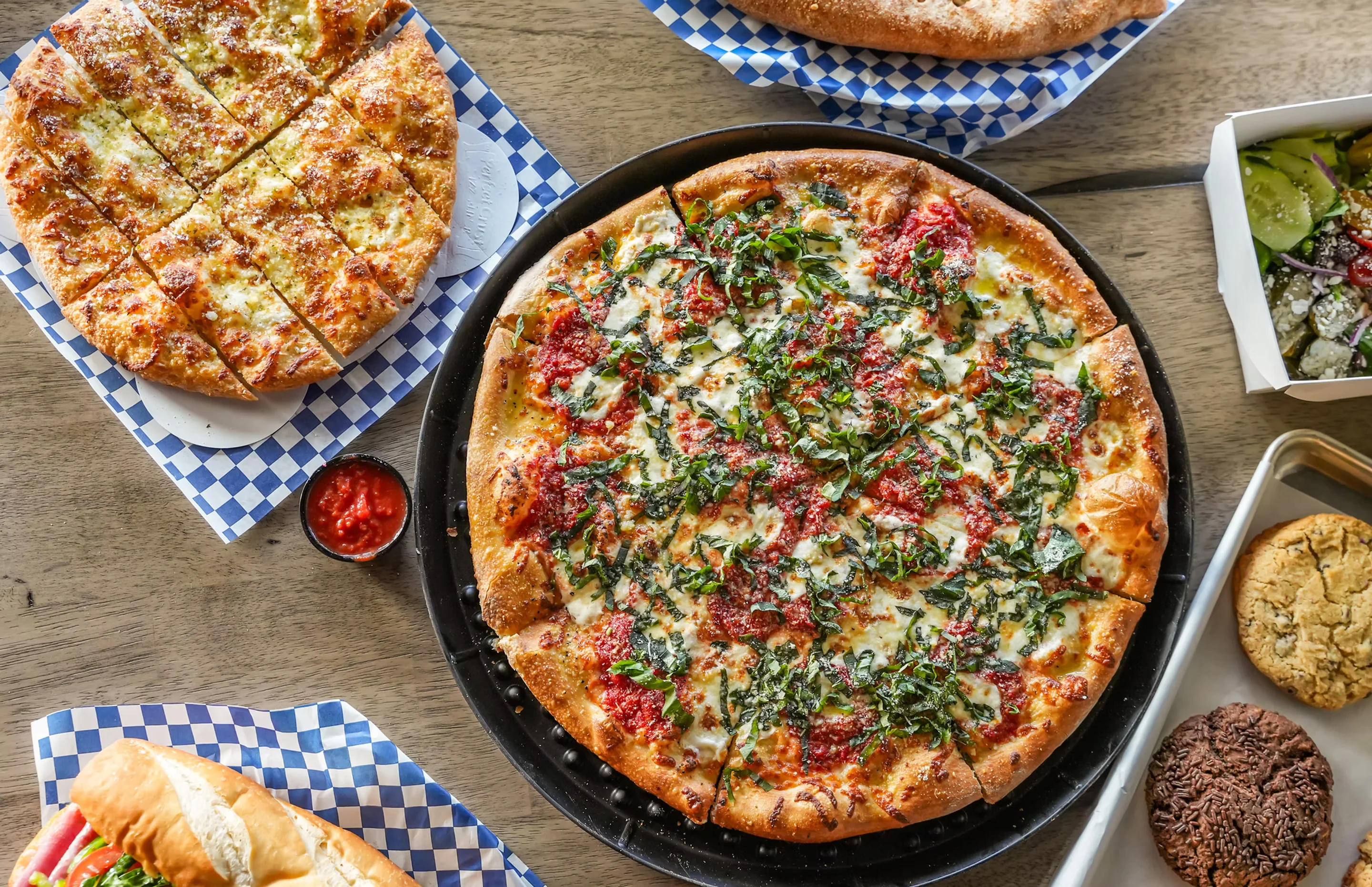 Goombas New York-style pizzas, grinder, calzone, salad, and giant cookies arranged on a table.