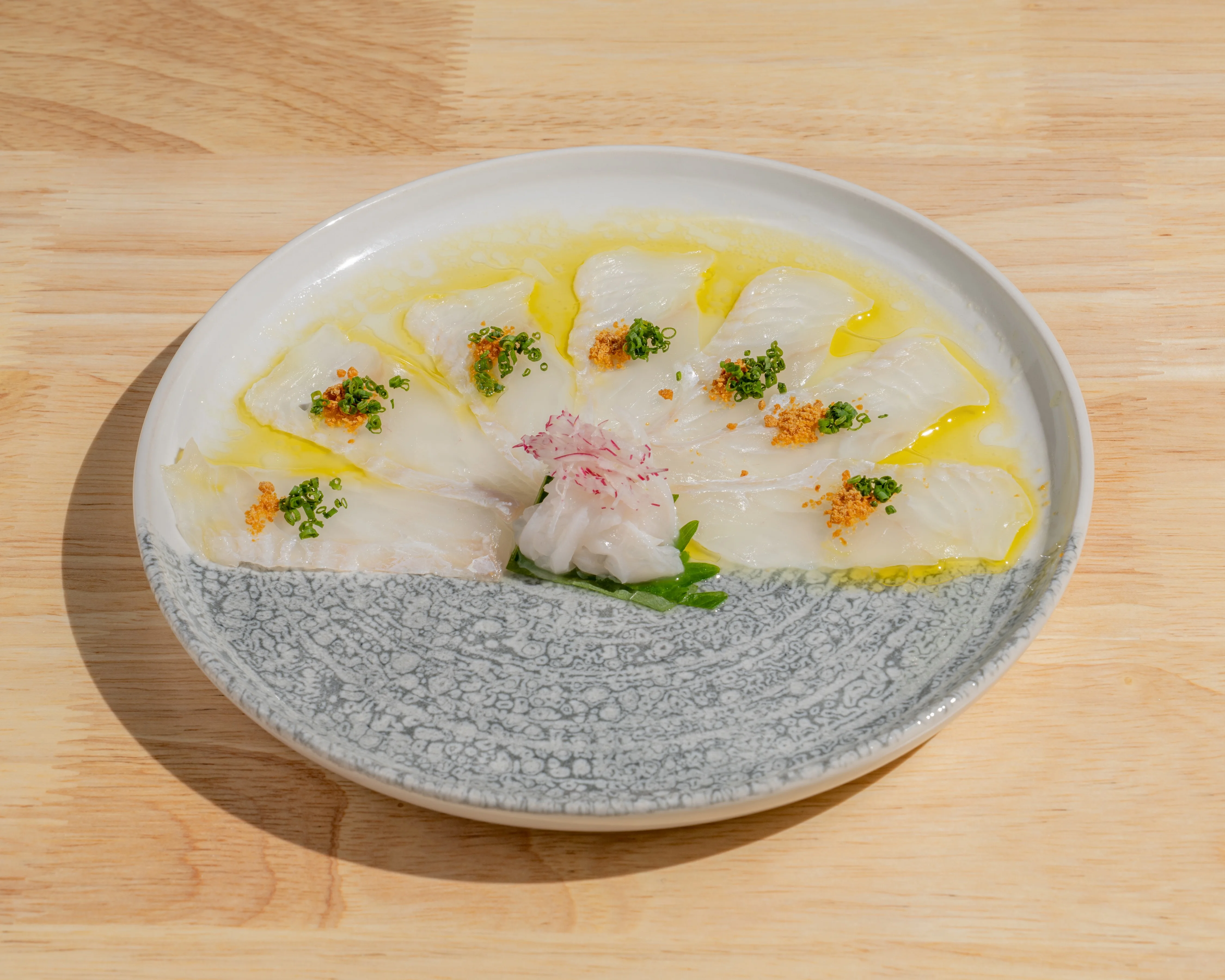 A plate of elegantly arranged thin slices of fish drizzled with olive oil and garnished with herbs.