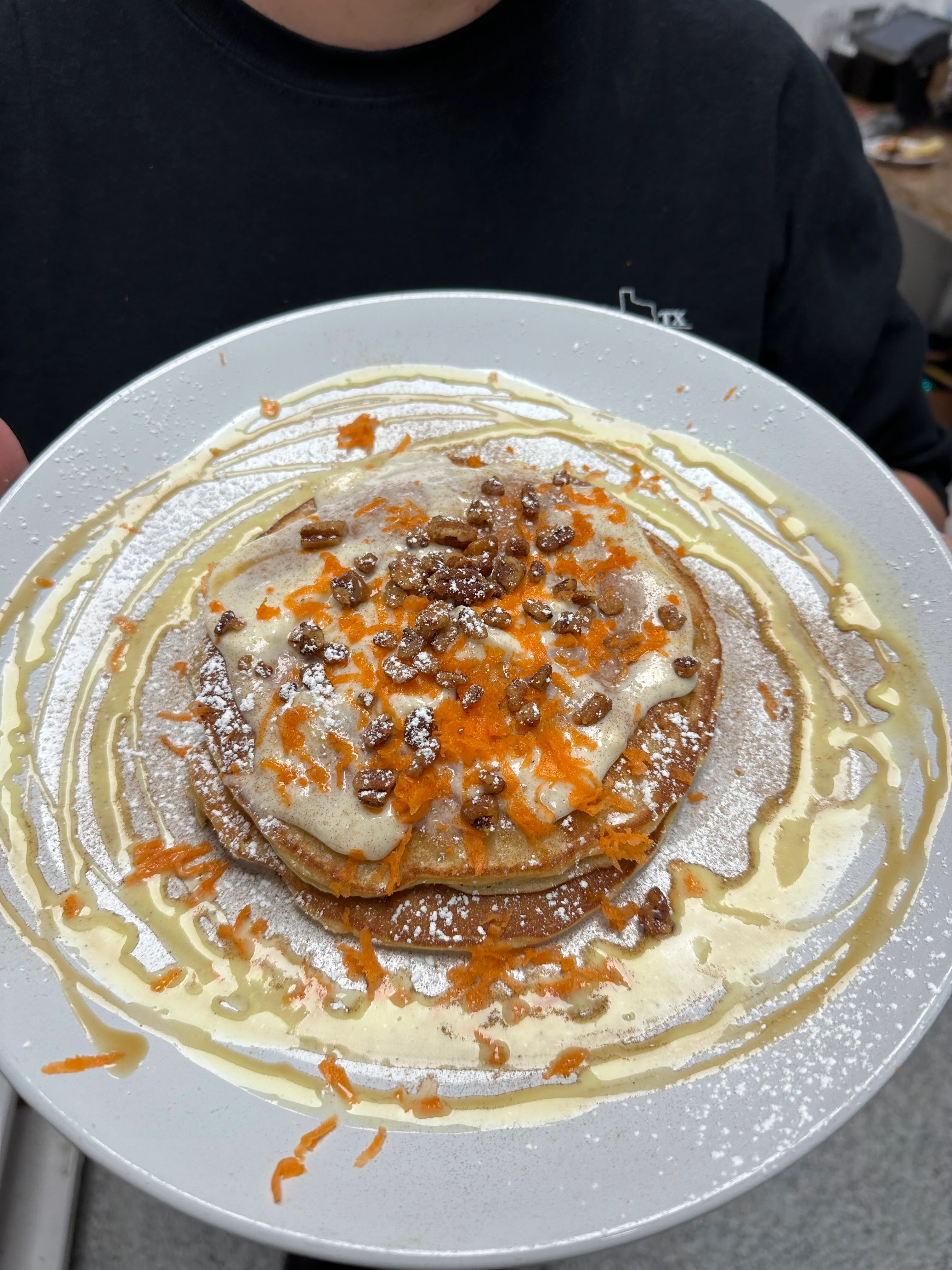 A plate of carrot cake pancakes topped with cream and sprinkled with chopped nuts and shredded carrots, drizzled with syrup.