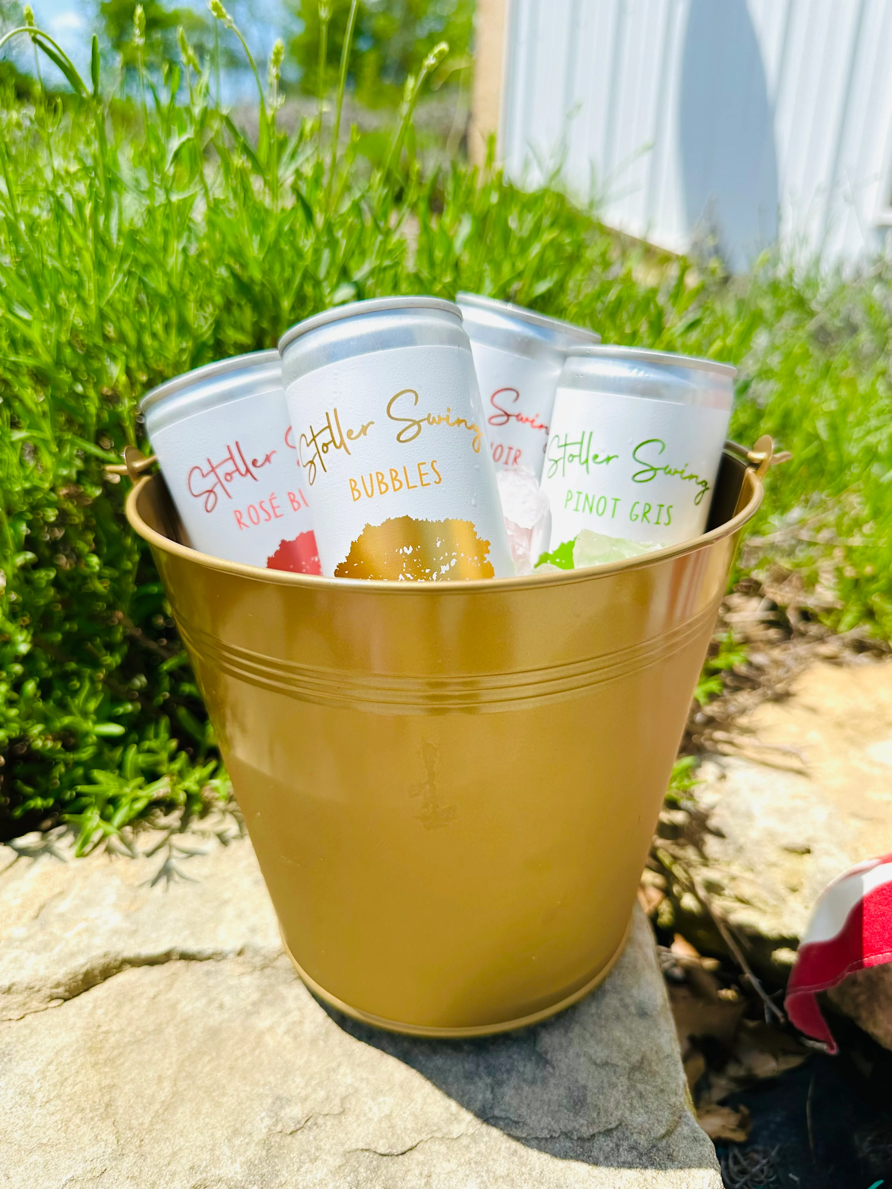 A golden bucket filled with various cans of wine, displayed on a stone surface with greenery in the background.