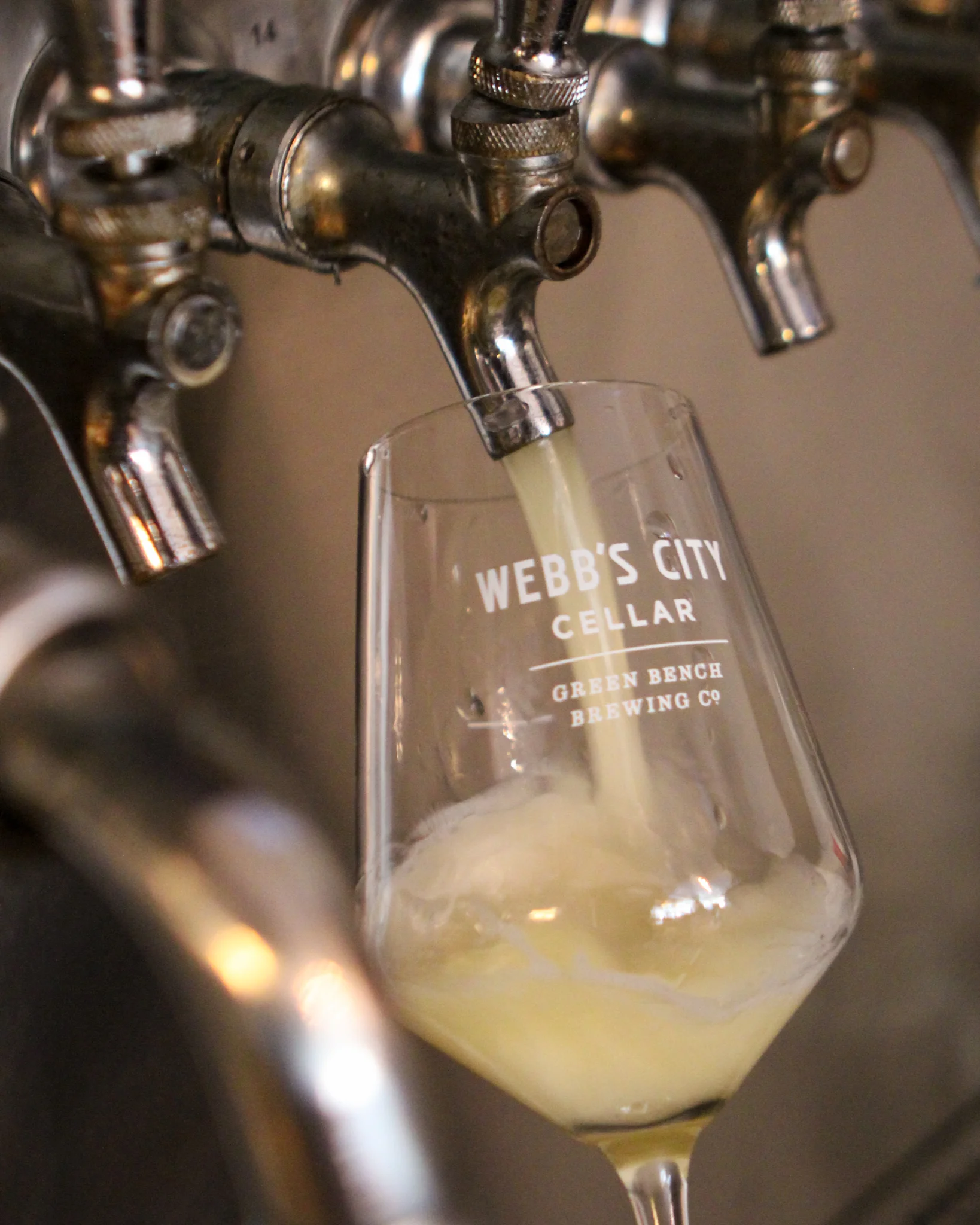 A glass being filled with a light-colored beverage from a metal tap at a brewery.