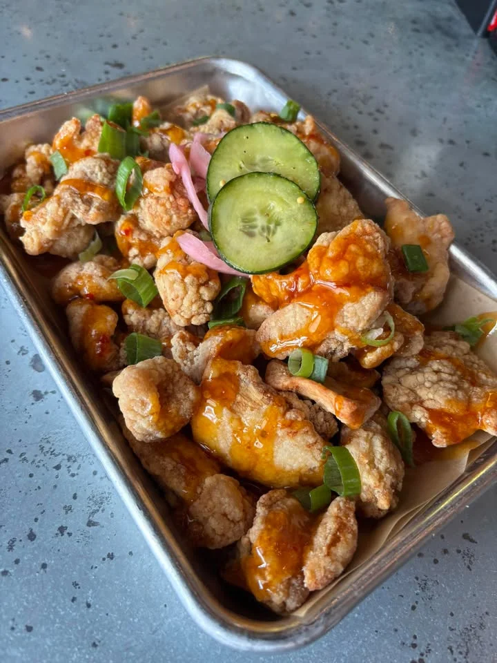 A tray filled with crispy pieces of chicken coated in a spicy sauce, garnished with sliced cucumbers and green onions.