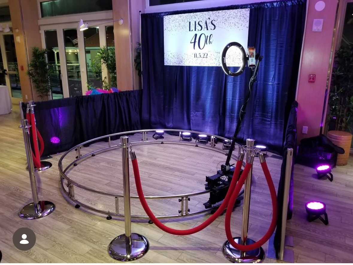 An event setup with a circular platform surrounded by red ropes, featuring a ring light for photography. The backdrop announces a 40th birthday celebration.