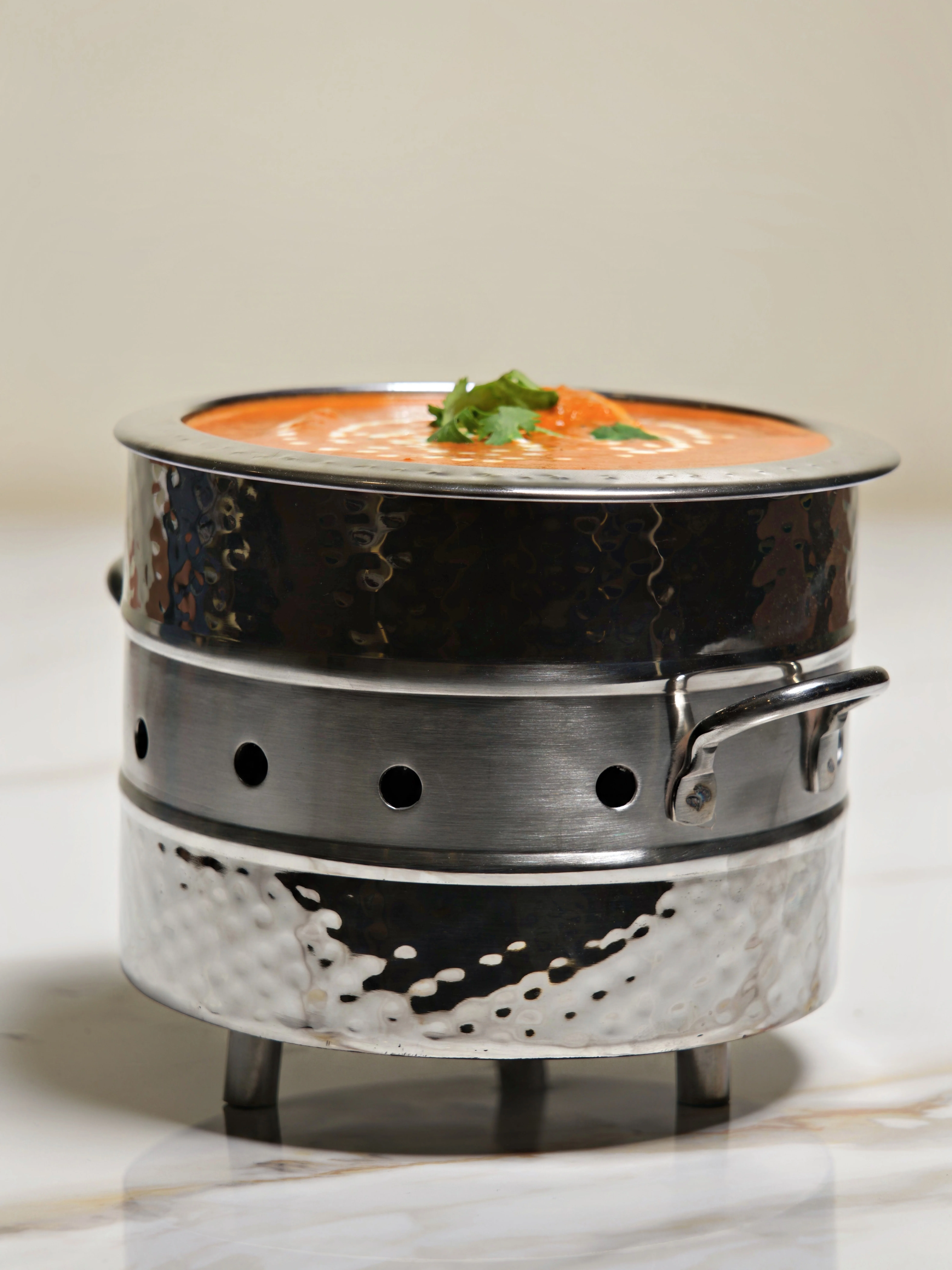 A traditional Indian dish served in a steamer pot, featuring a creamy orange sauce, decorated with swirls of cream and fresh herbs on top.