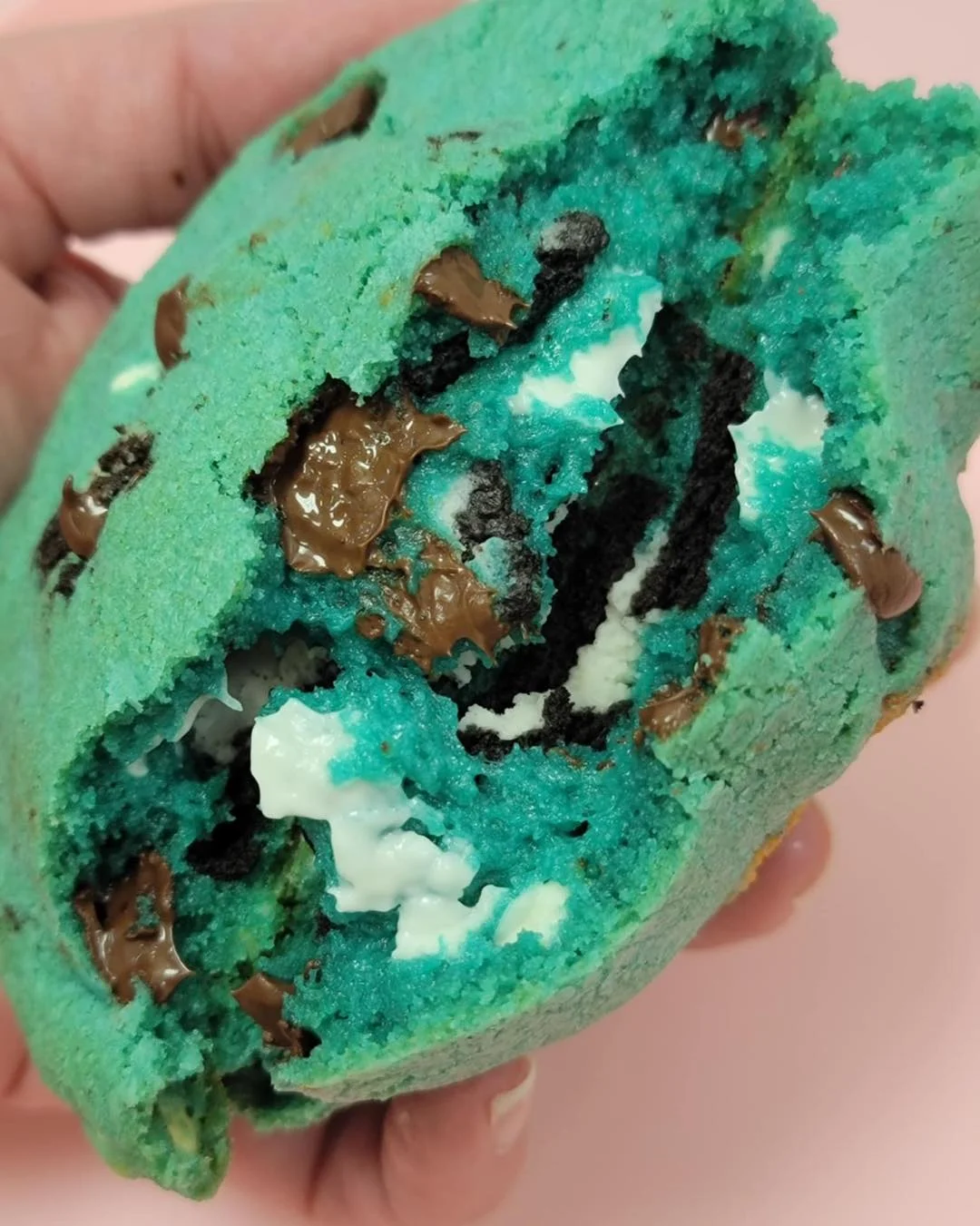 A large blue stuffed cookie broken open, filled with Oreo pieces, white cream, and melted chocolate chips.