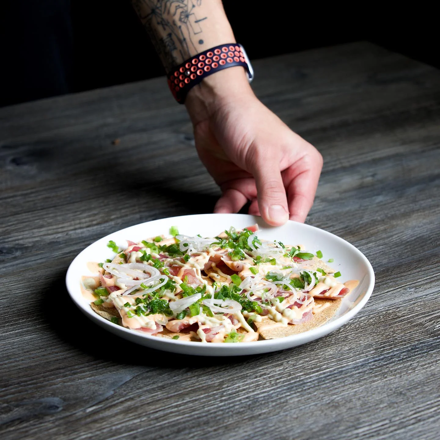 A hand placing a white plate filled with tuna nachos onto a grey wooden table, showcasing crispy tortilla chips layered with thin slices of seared tuna, drizzled with spicy mayo and white sauce, and topped with sliced onions and chopped green scallions.