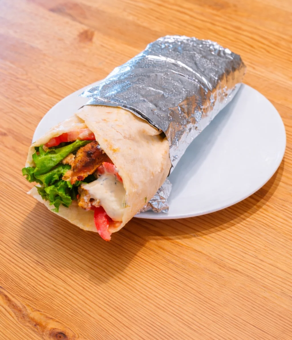 A wrapped sandwich made with pita bread, filled with grilled meat, lettuce, and tomatoes, resting on a plate.