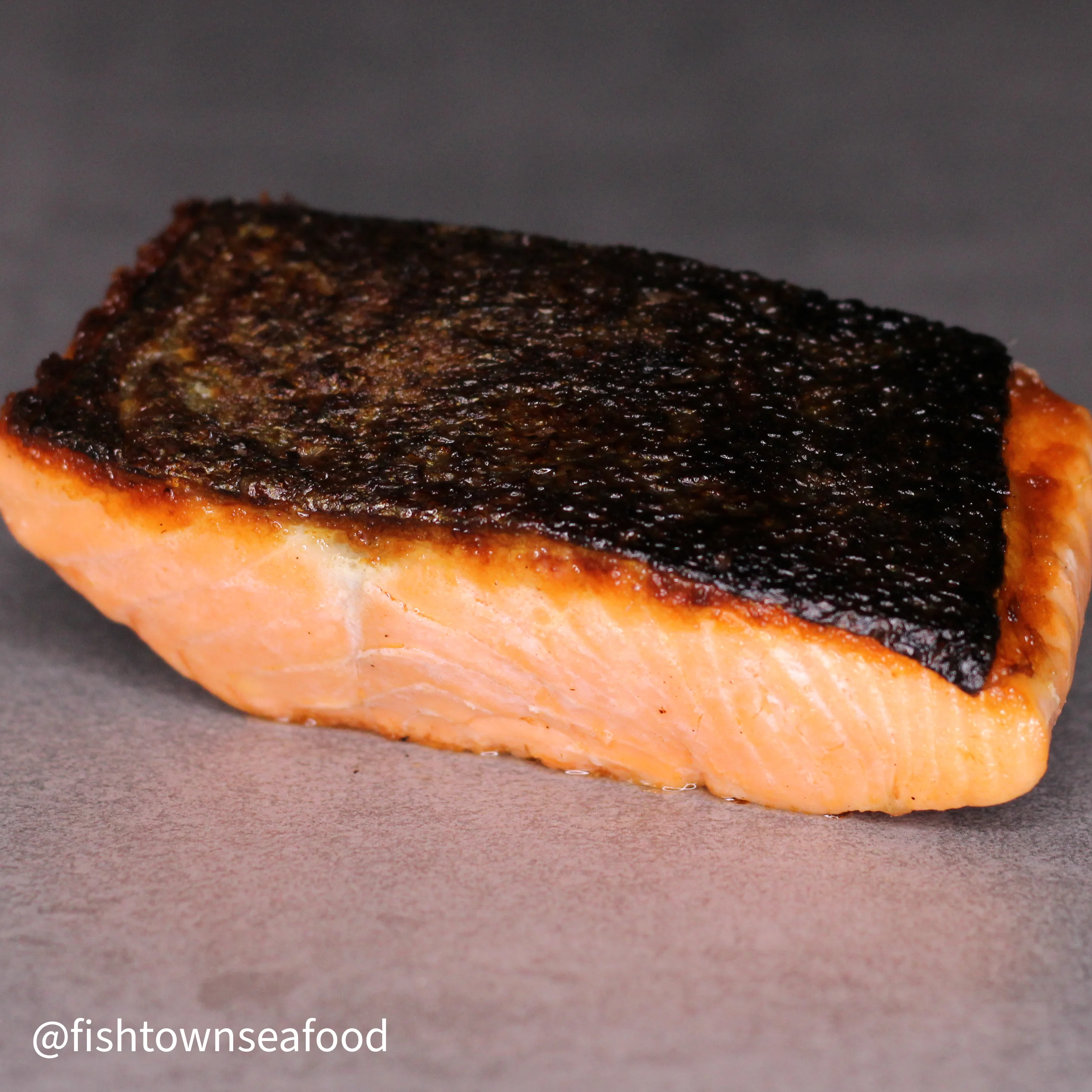 A piece of cooked salmon, featuring a crispy skin on top and a tender interior, placed on a neutral background.