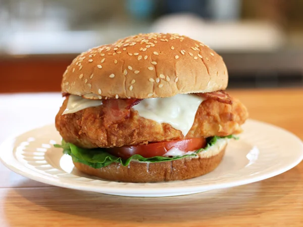 Delicious bacon cheddar melt crispy chicken sandwich from Countryside Drive-In, featuring golden fried chicken, melted cheddar cheese, and crispy bacon on a sesame-seeded bun. Served with crispy fries and a cold drink on a rustic wooden table, capturing our signature hearty meal.