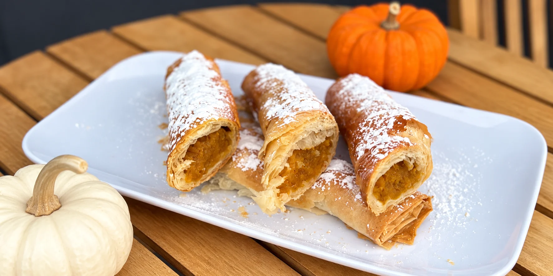 A plate featuring rolled pastries filled with pumpkin, dusted with powdered sugar, accompanied by small decorative pumpkins.