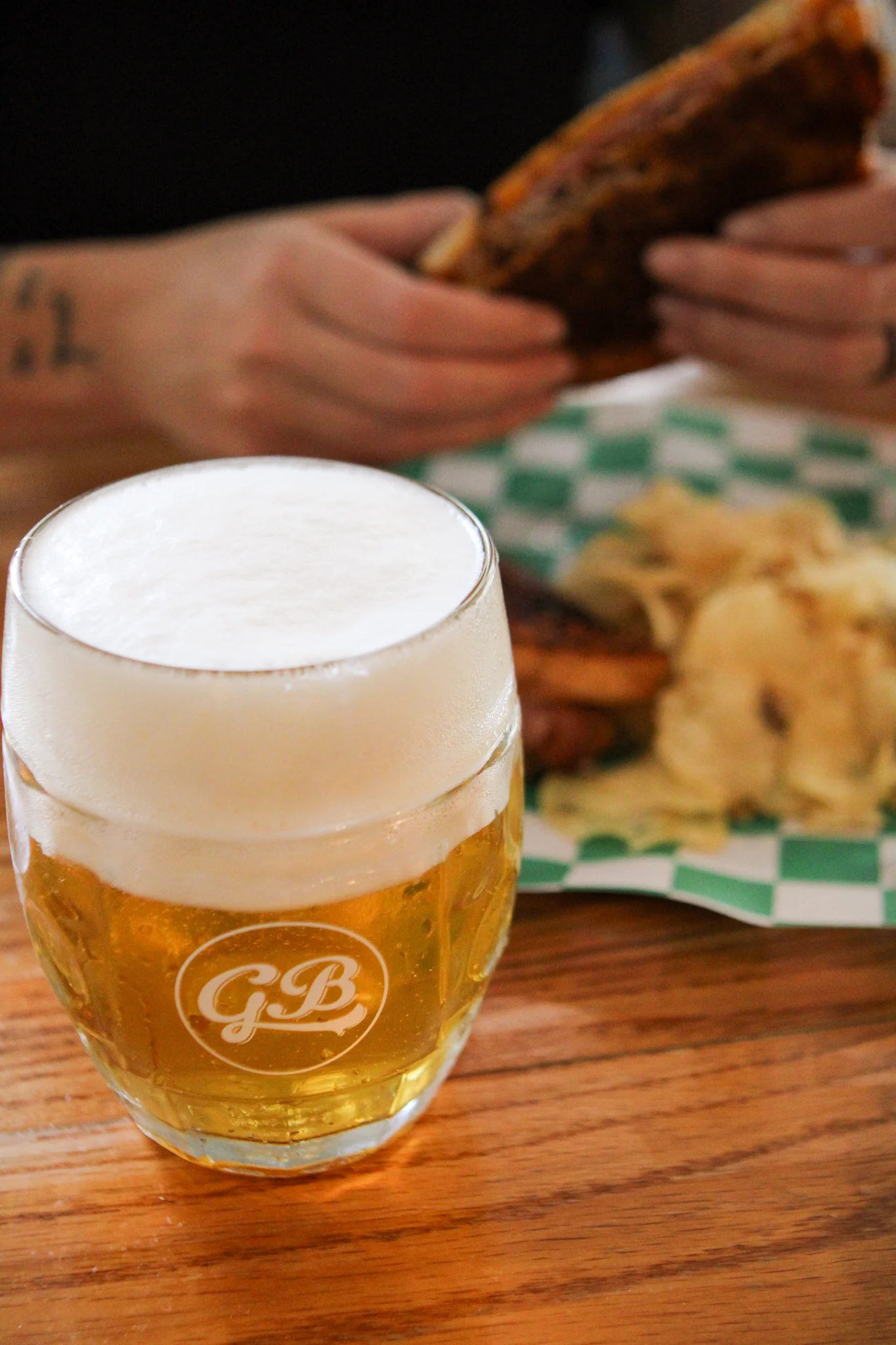 A close-up of a beer glass filled with foamy beer, placed on a wooden table, while a person holds a sandwich in the background.