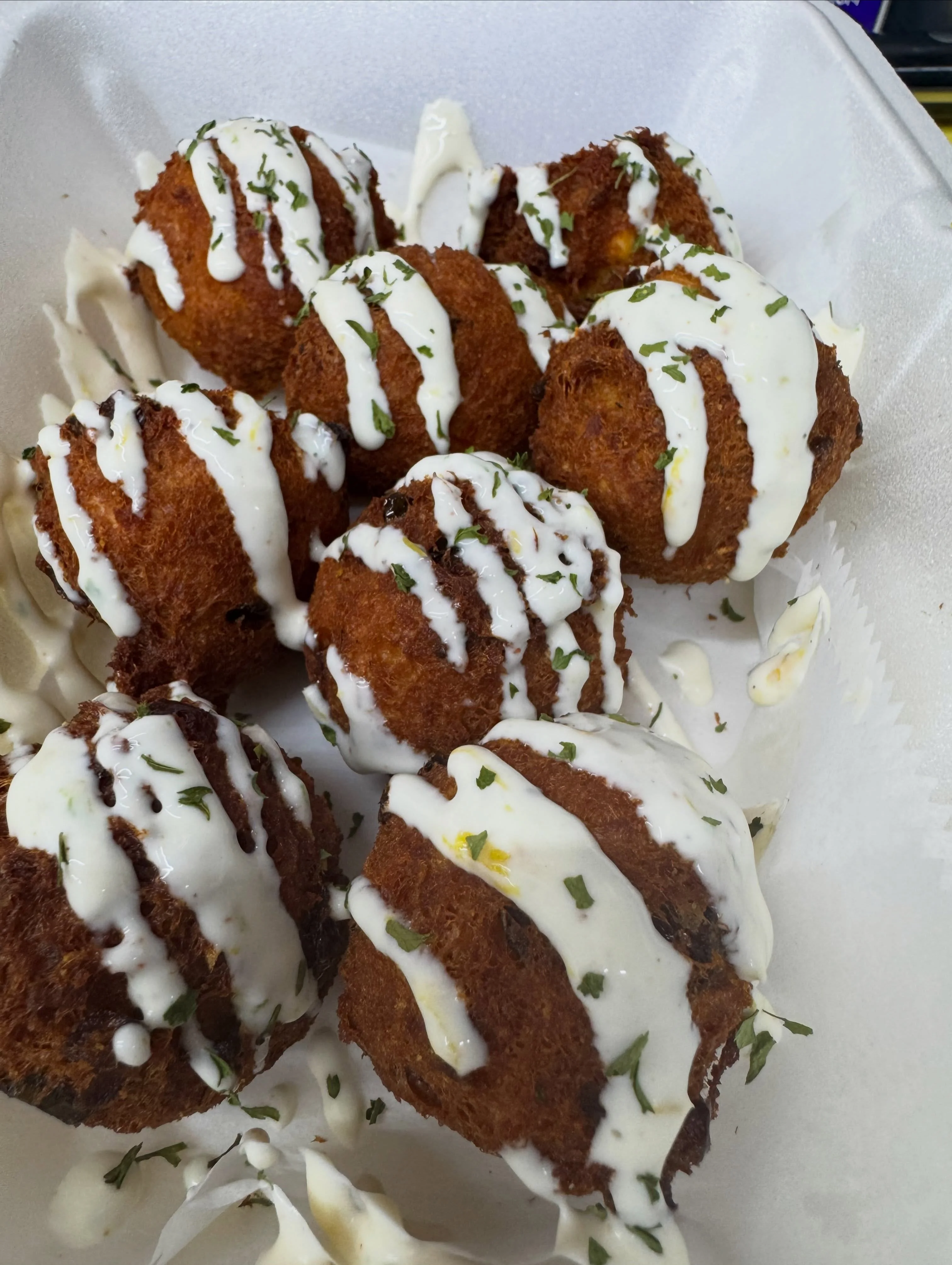 A container filled with golden fried spherical bites, topped with a creamy sauce and garnished with herbs.