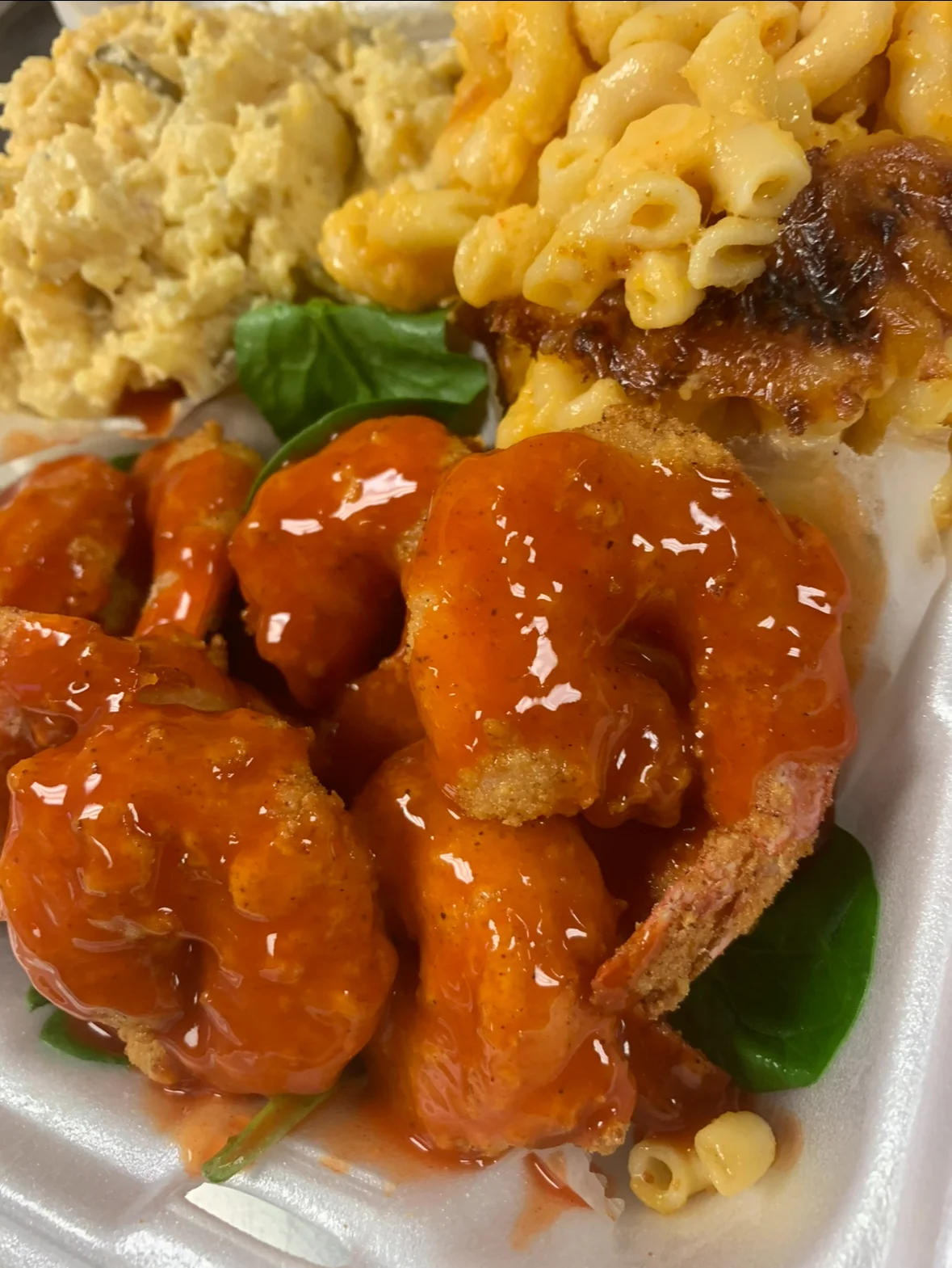 A serving of fried shrimp coated in a spicy sauce, accompanied by macaroni and cheese, coleslaw, and fresh greens.