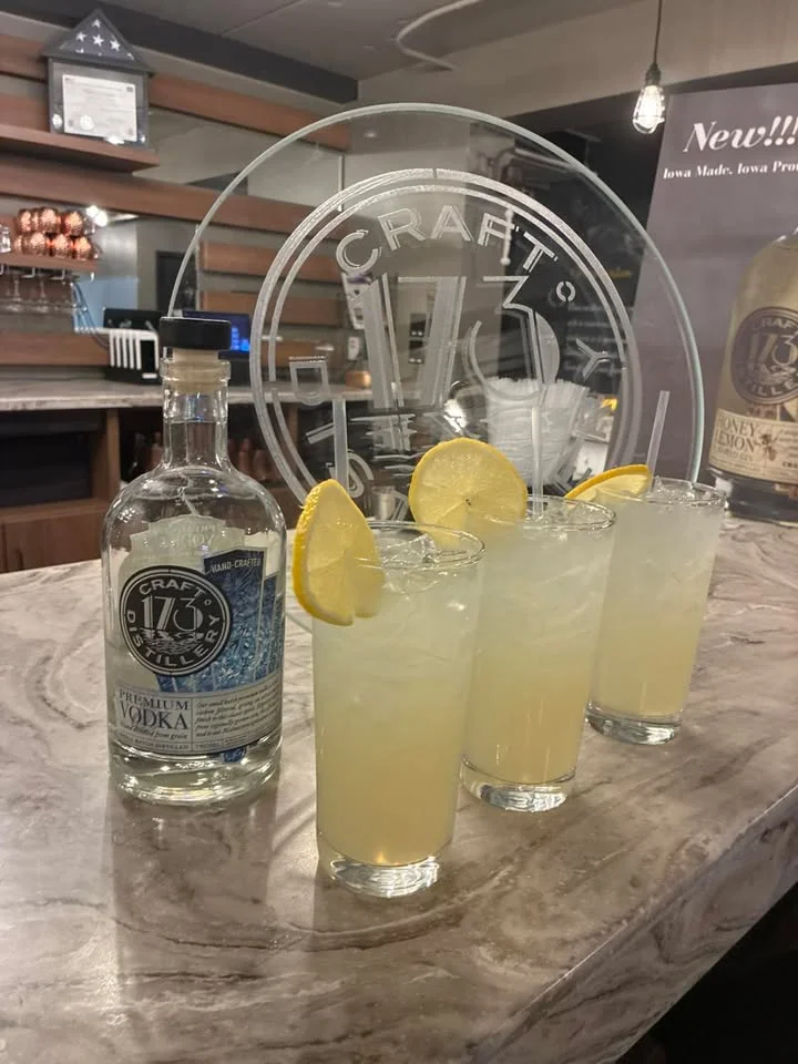 Four drinks are served alongside a bottle of vodka. Each drink is garnished with a lemon slice, presented on a polished surface.