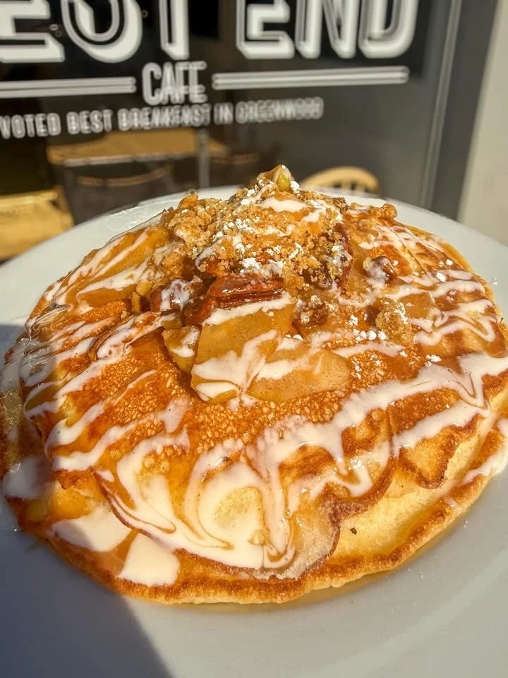 A stack of fluffy pancakes drizzled with creamy icing and topped with apple pieces and a sprinkle of powdered sugar.
